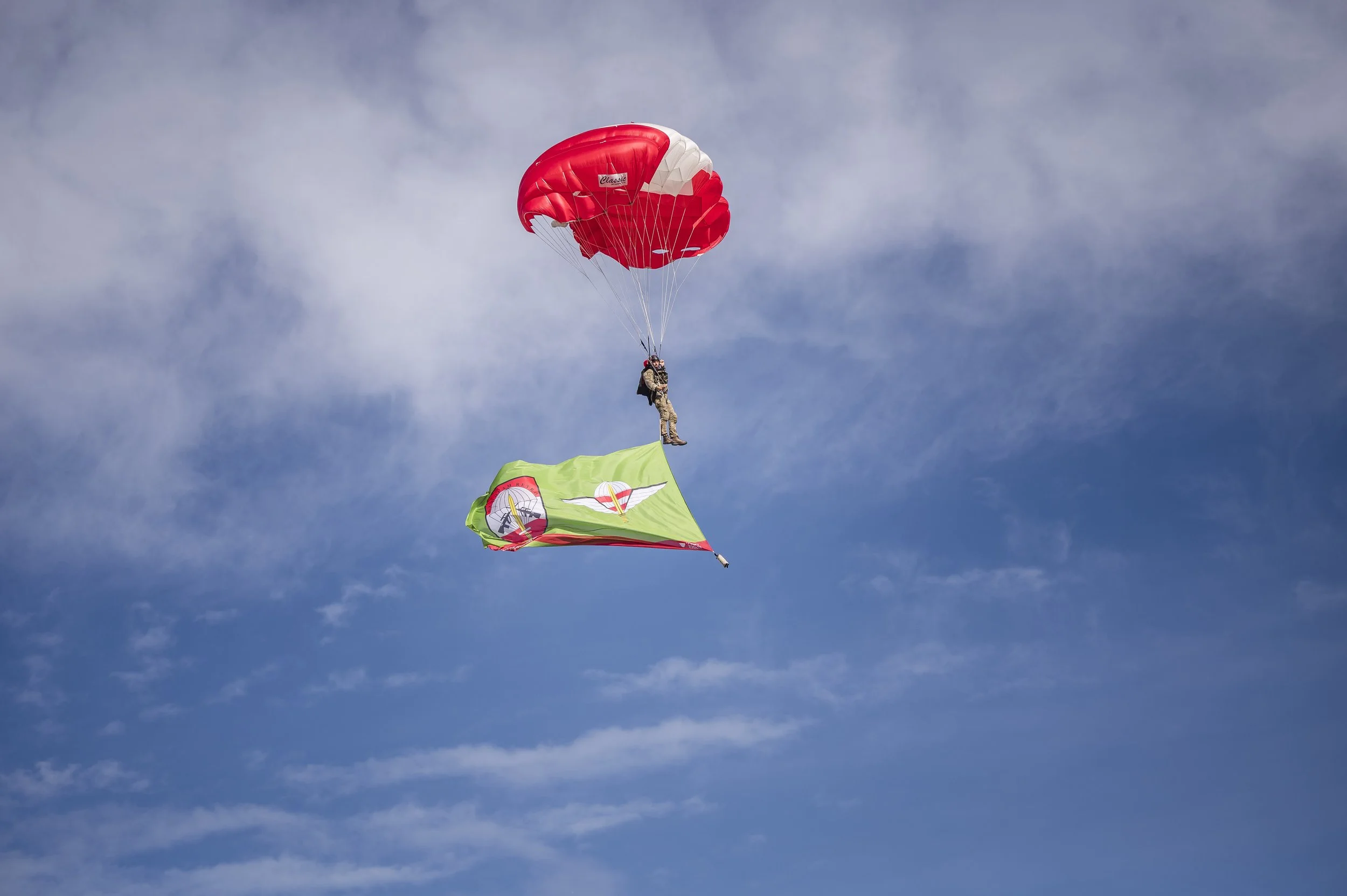 Ein Fallschirmspringer, der mit einem roten und weißen Fallschirm fliegt und eine grüne Flagge hält, während er durch den Himmel über Wolken fliegt.