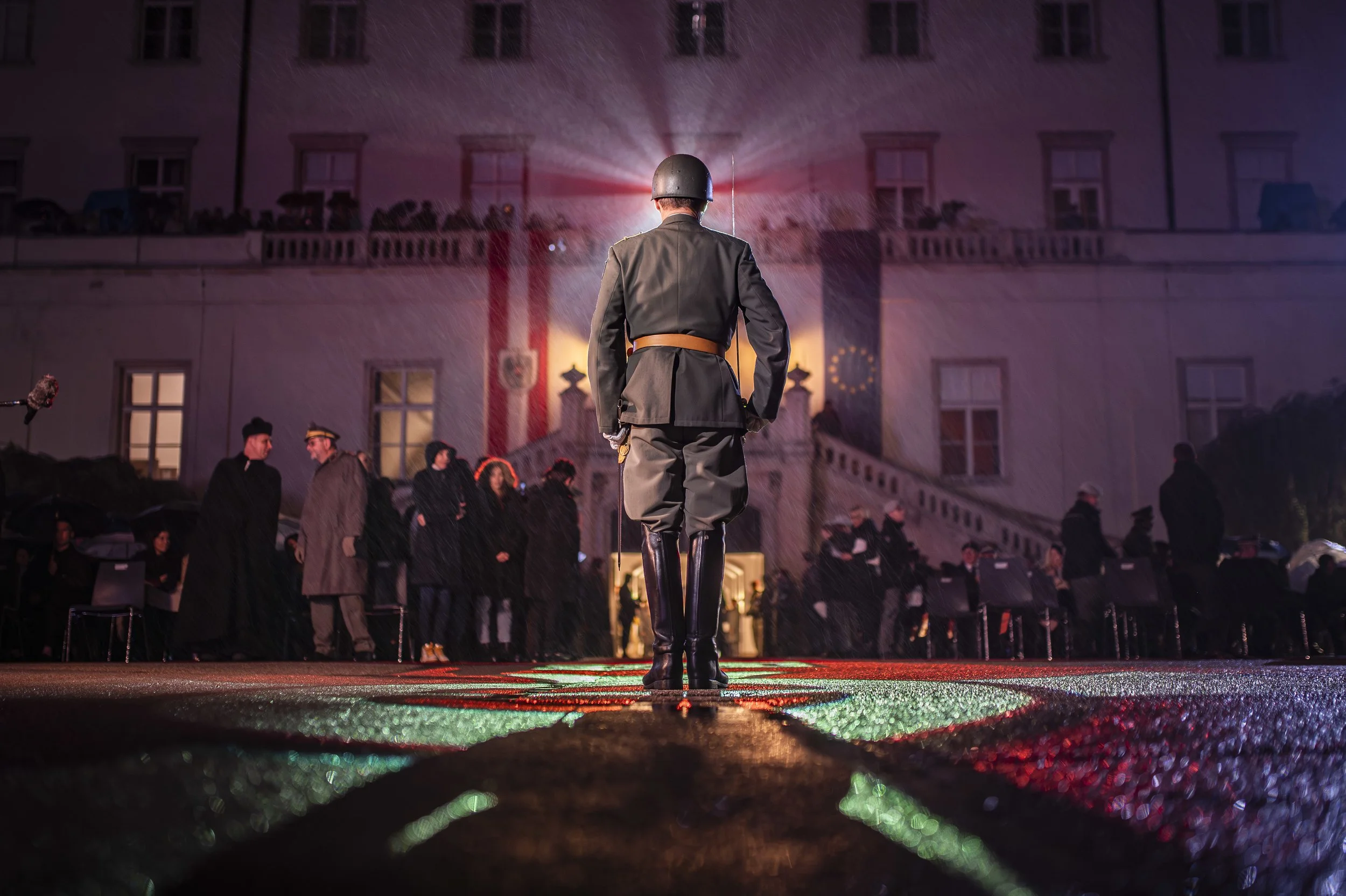 Ein Soldat in Uniform und Helm steht auf einer nassen Straße, im Hintergrund ein Gebäude mit mehreren Fenstern und Menschen, die draußen stehen, bei Nacht. Es regnet, und die Straße ist mit bunten Lichterreflexen bedeckt.