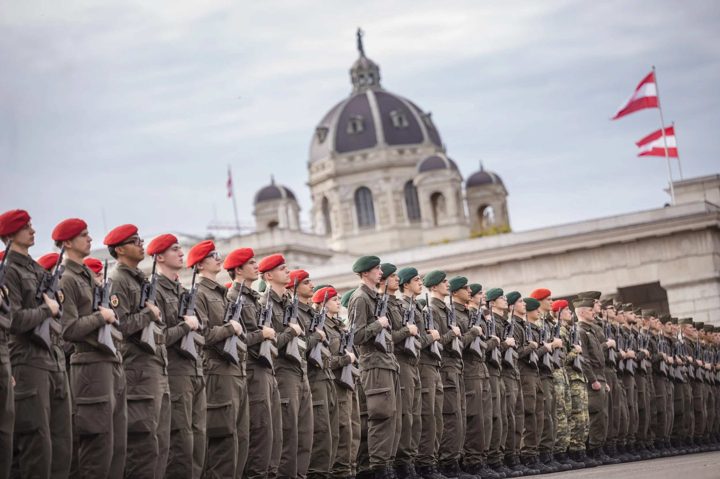 Soldaten in Formation mit roten und grünen Hüten stehen in einer Reihe vor einer historischen Gebäudefront, mit Flaggen im Hintergrund.