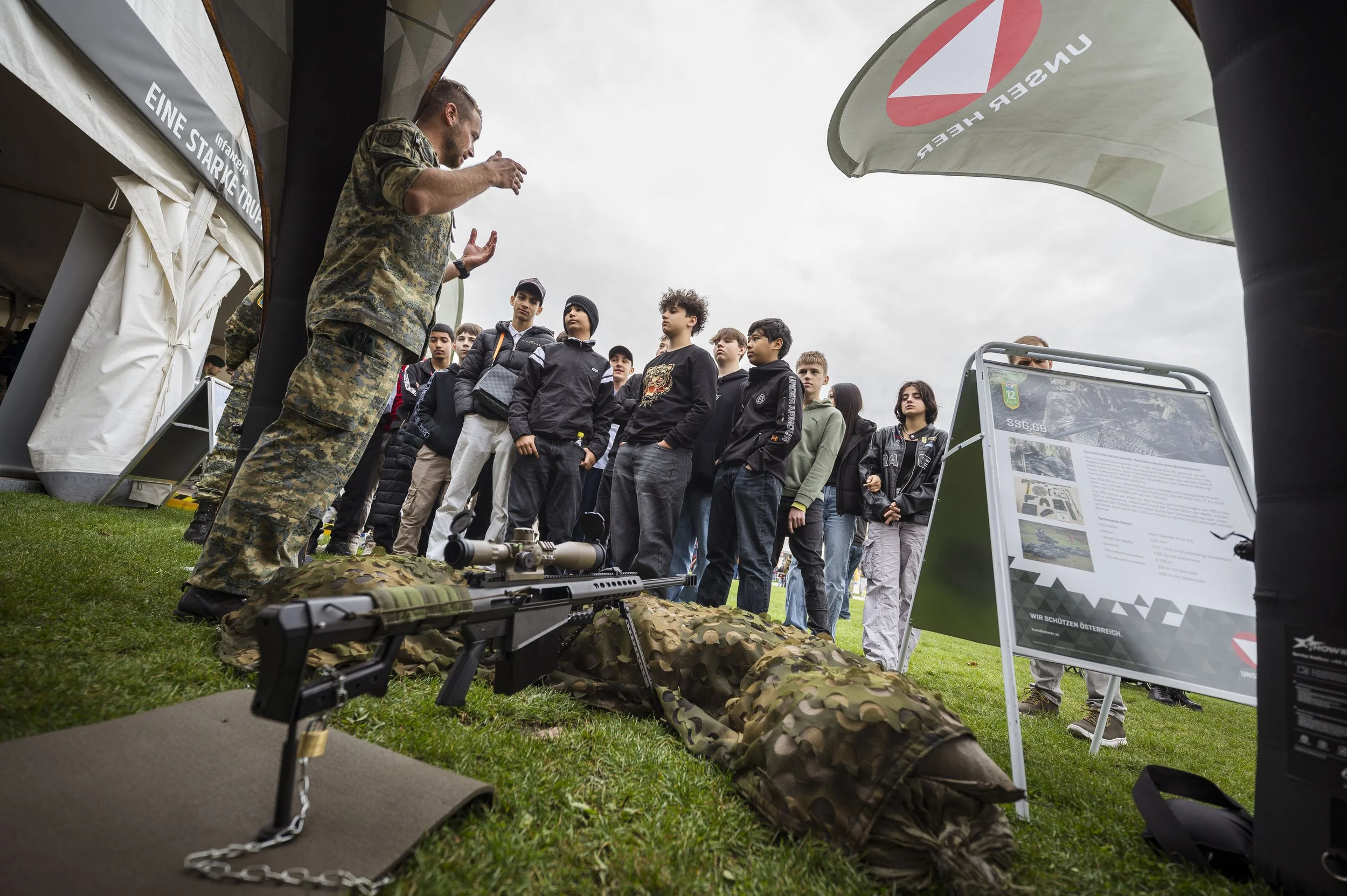 Ein Militärs erklärt einer Gruppe von Jugendlichen an einem Outdoor-Informationsstand Waffen und Ausrüstung. Im Vordergrund liegt eine Waffe auf einer camouflierten Decke, im Hintergrund sind Jugendliche, ein Poster mit Text und Fahnen. Das Wetter is