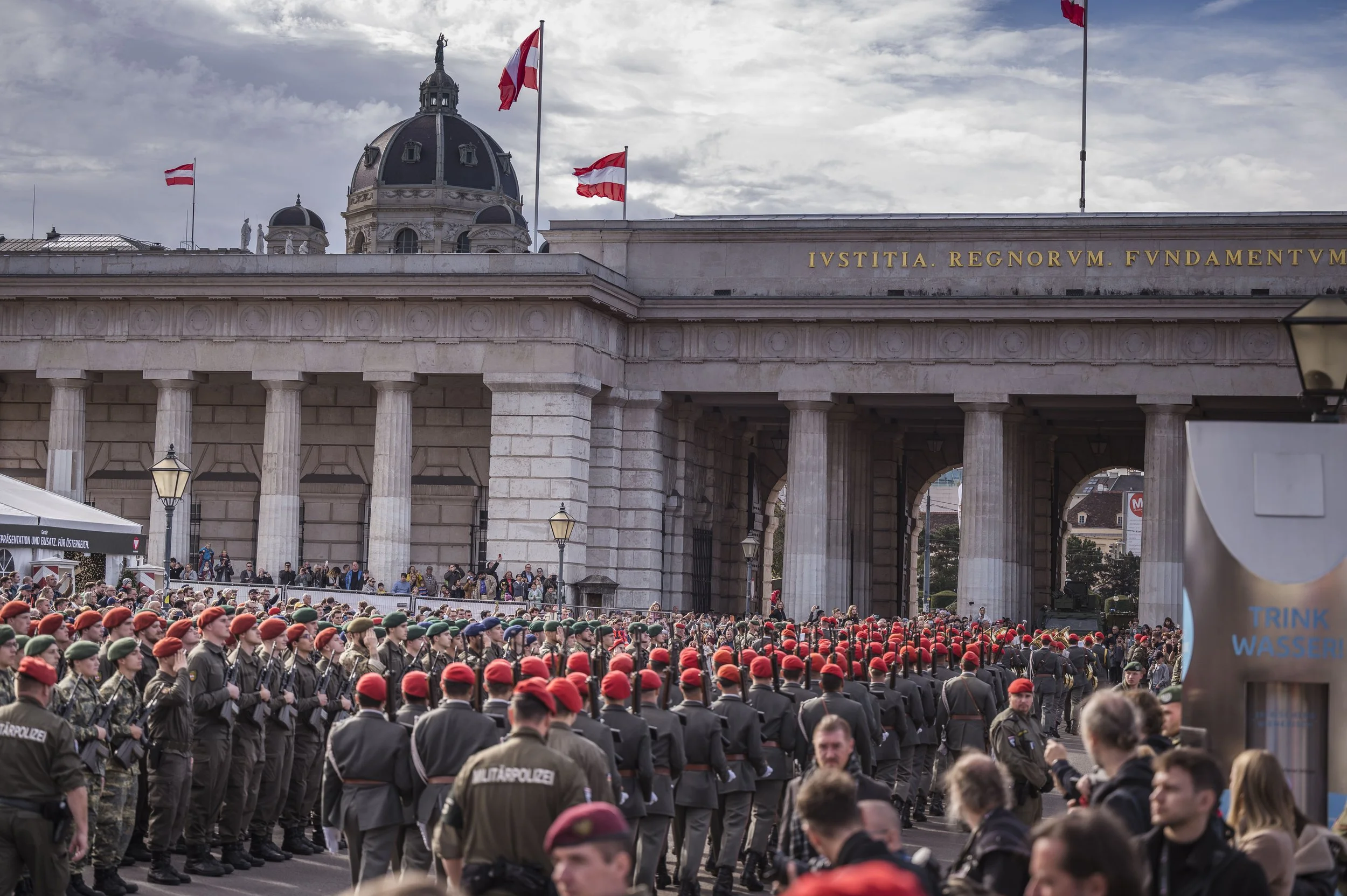 Militärparade mit Soldaten in Uniformen und roten Helmen in Berlin, Deutschland, vor dem Brandenburger Tor mit Flaggen und Menschenmengen im Hintergrund.