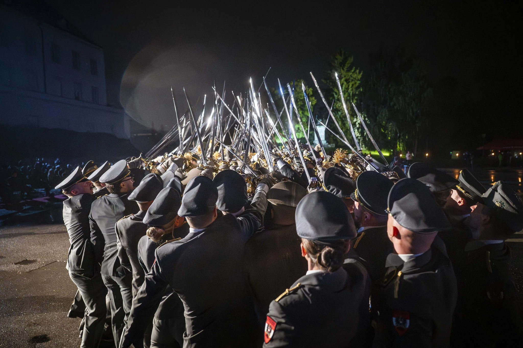 Truppsoldaten in Uniformen, die im Gleichschritt marschieren, mit angehobenen Säbeln. Es ist nachts und im Hintergrund sind Bäume, ein Gebäude sowie einige Lichter sichtbar.