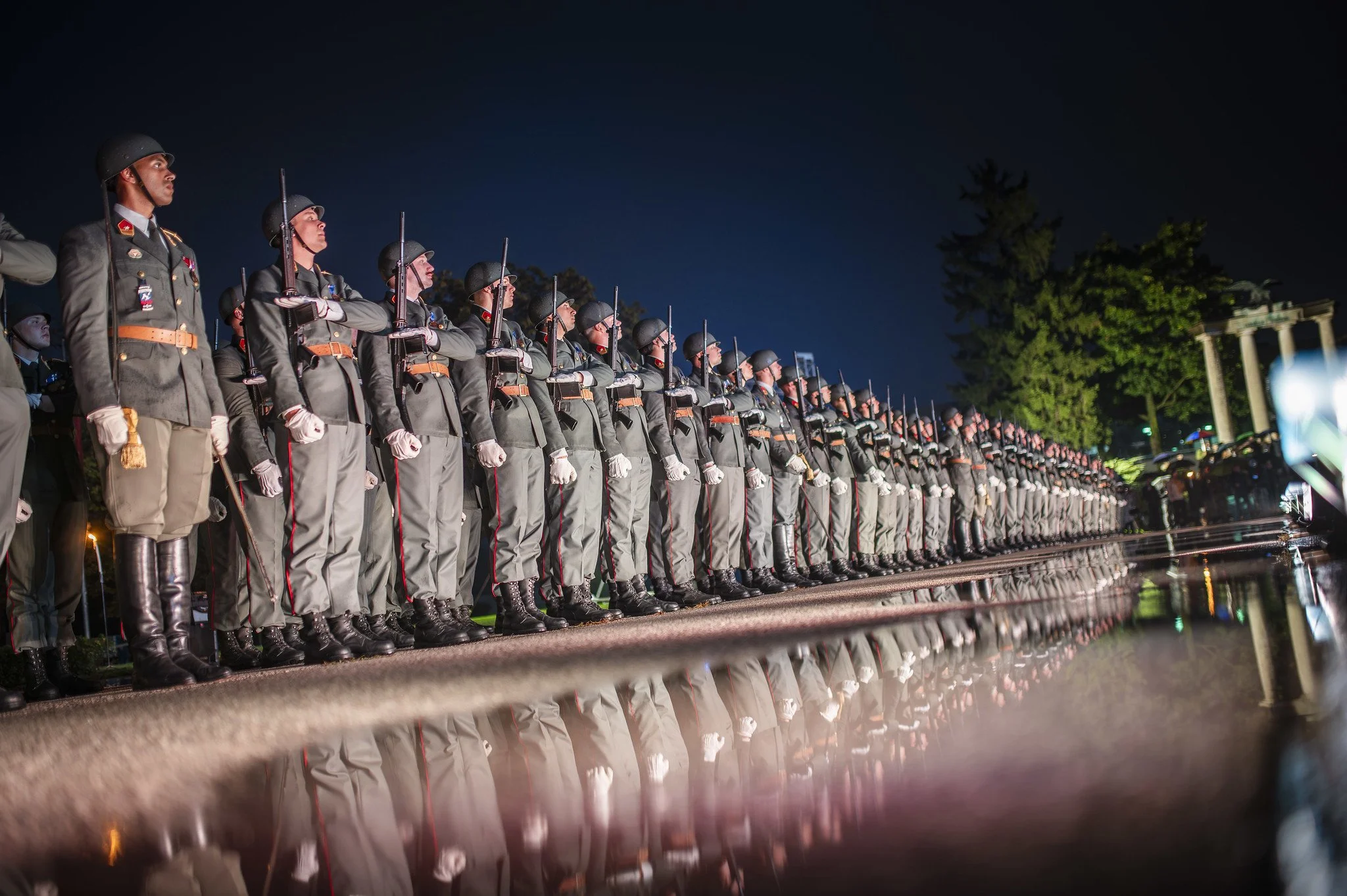 Reihung von Soldaten in Uniform bei einer Zeremonie bei Nacht, reflektierende Wasseroberfläche im Vordergrund, Bäume und Säulen im Hintergrund.