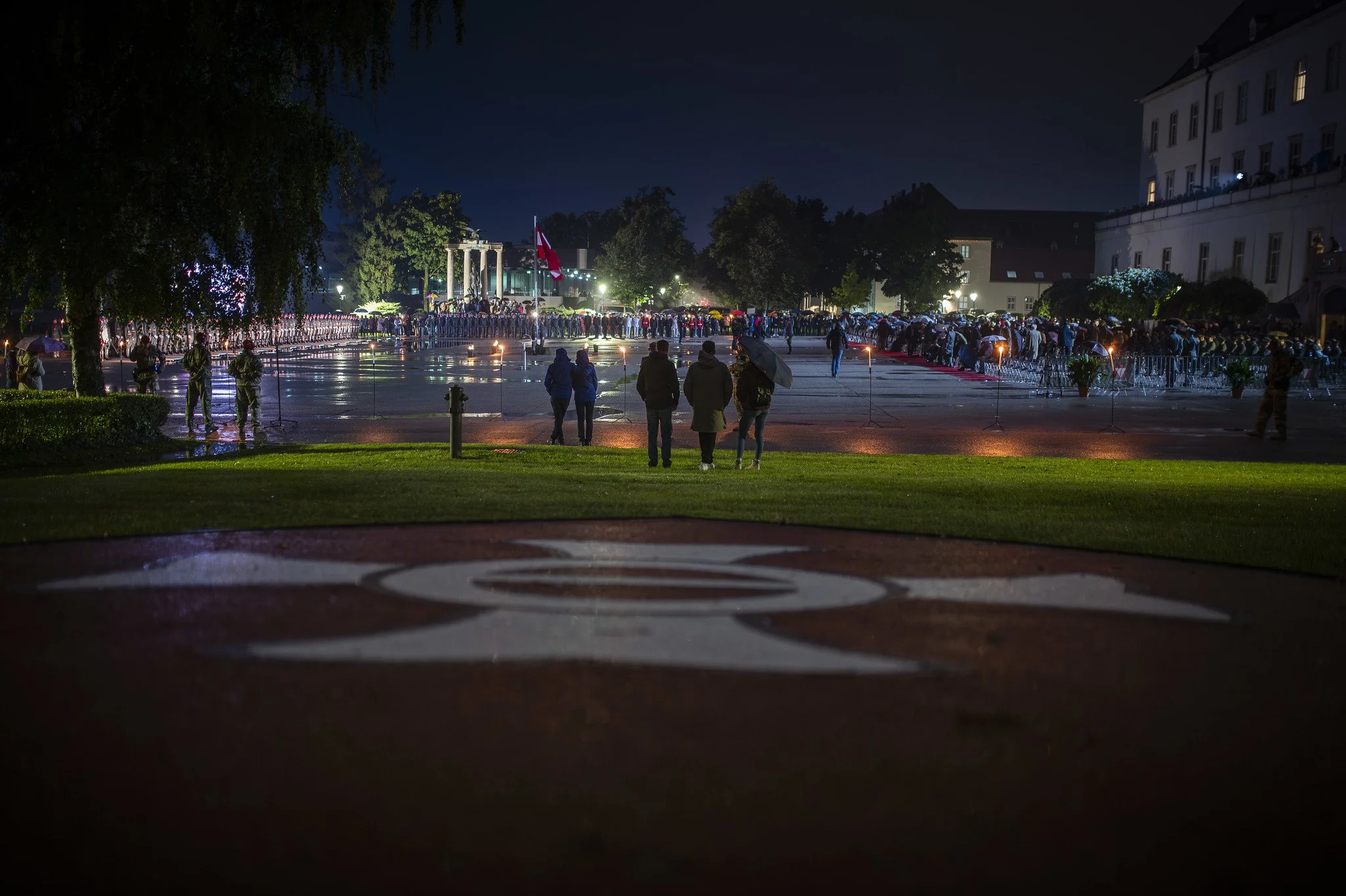 Menschen versammeln sich bei Nacht auf einem Platz, einige mit Regenschirmen, es scheint eine Warteschlange oder eine Zeremonie zu geben, mit einem Flagge in der Mitte und Toren oder Säulen im Hintergrund, unterbewölktem Himmel