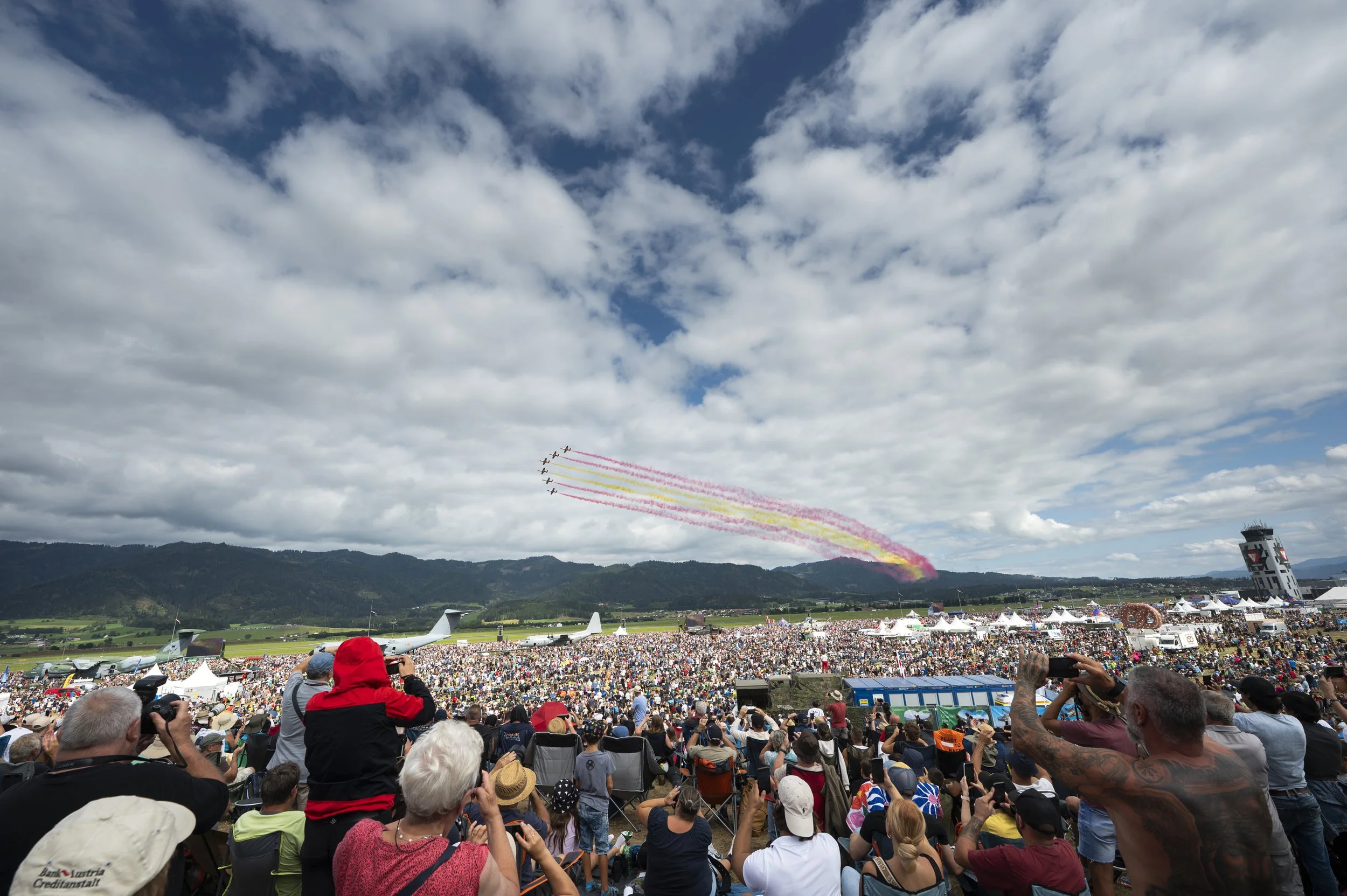 Menschenmenge bei Flugshow mit Flugzeugen, die bunte Rauchfahnen am Himmel hinterlassen, in einer ländlichen Gegend mit Bergen im Hintergrund.