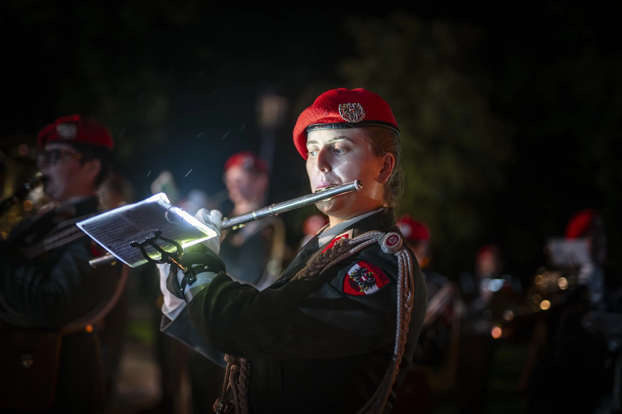 Musikerin in Uniform mit roter Mütze spielt Flöte bei Nacht, umgeben von anderen Musikern, im Hintergrund dunkle Umgebung.