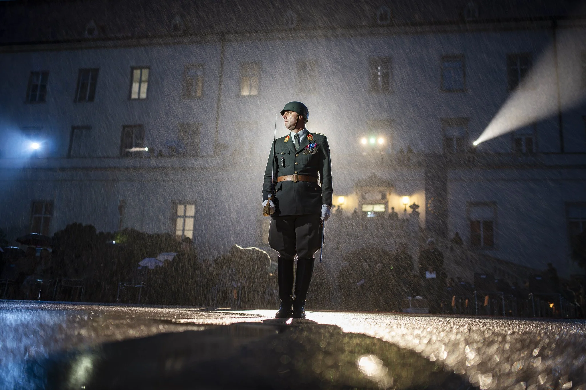 Ein Soldat steht in Uniform bei Nacht im Regen, mit Scheinwerfer beleuchtet, vor einem dunklen Gebäude.