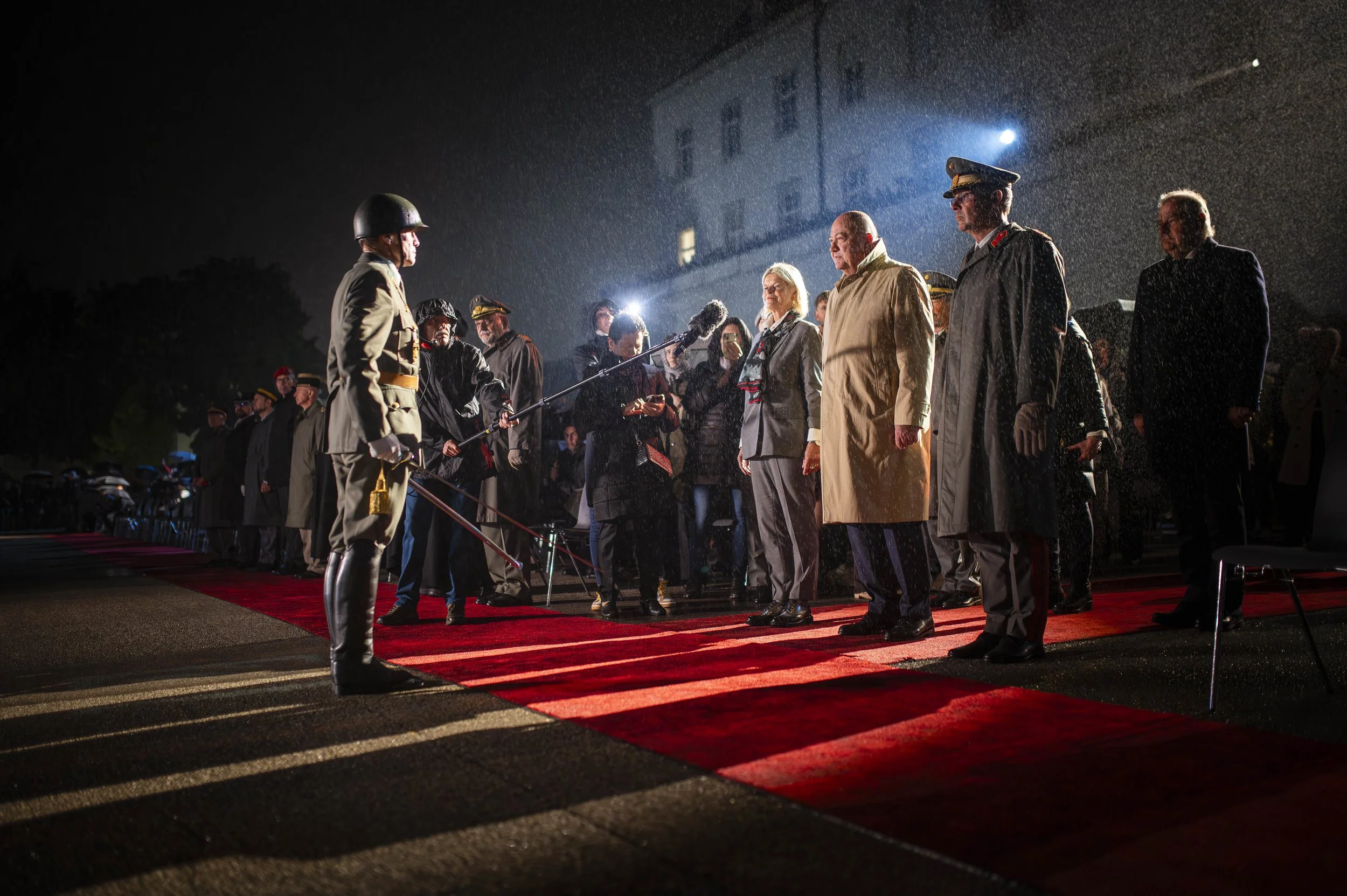 Militärische Zeremonie bei Nacht mit Soldaten, hochrangigen Persönlichkeiten und Medien, die auf einem roten Teppich stehen, während Regen fällt.