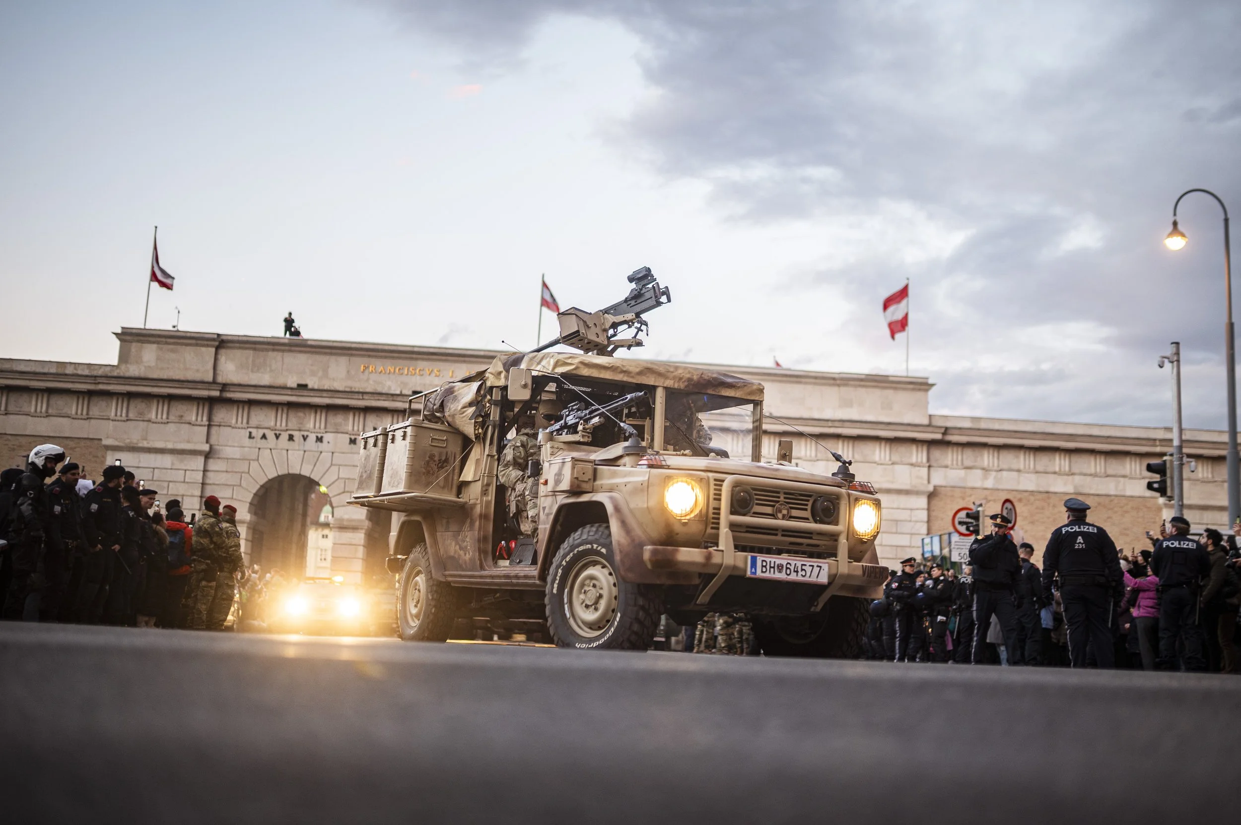 Ein Militärfahrzeug fährt durch eine Menschenmenge in einer Stadt, wahrscheinlich bei einer Demonstration oder Parade, bei Dämmerung oder Sonnenuntergang, mit Gebäuden, Menschen und Polizisten im Hintergrund.