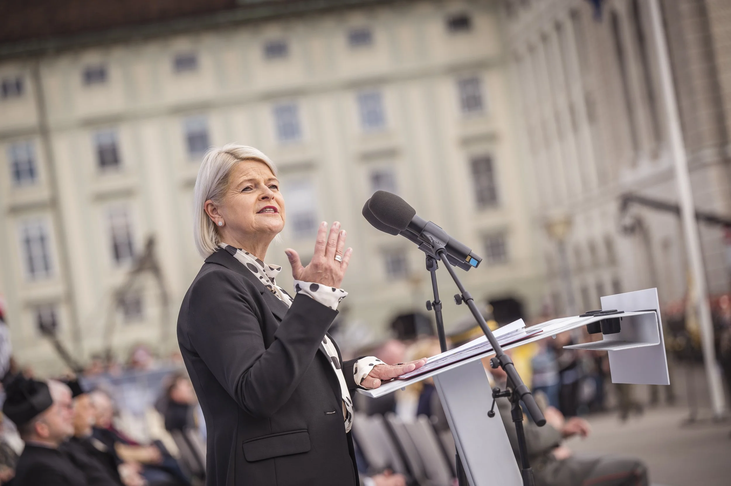 Eine älteren Frau beim öffentlichen Reden, im Hintergrund ein Gebäude mit mehreren Fenstern, sie trägt einen schwarzen Blazer und eine hell gepunktete Bluse, während sie vor einem Mikrofon spricht.