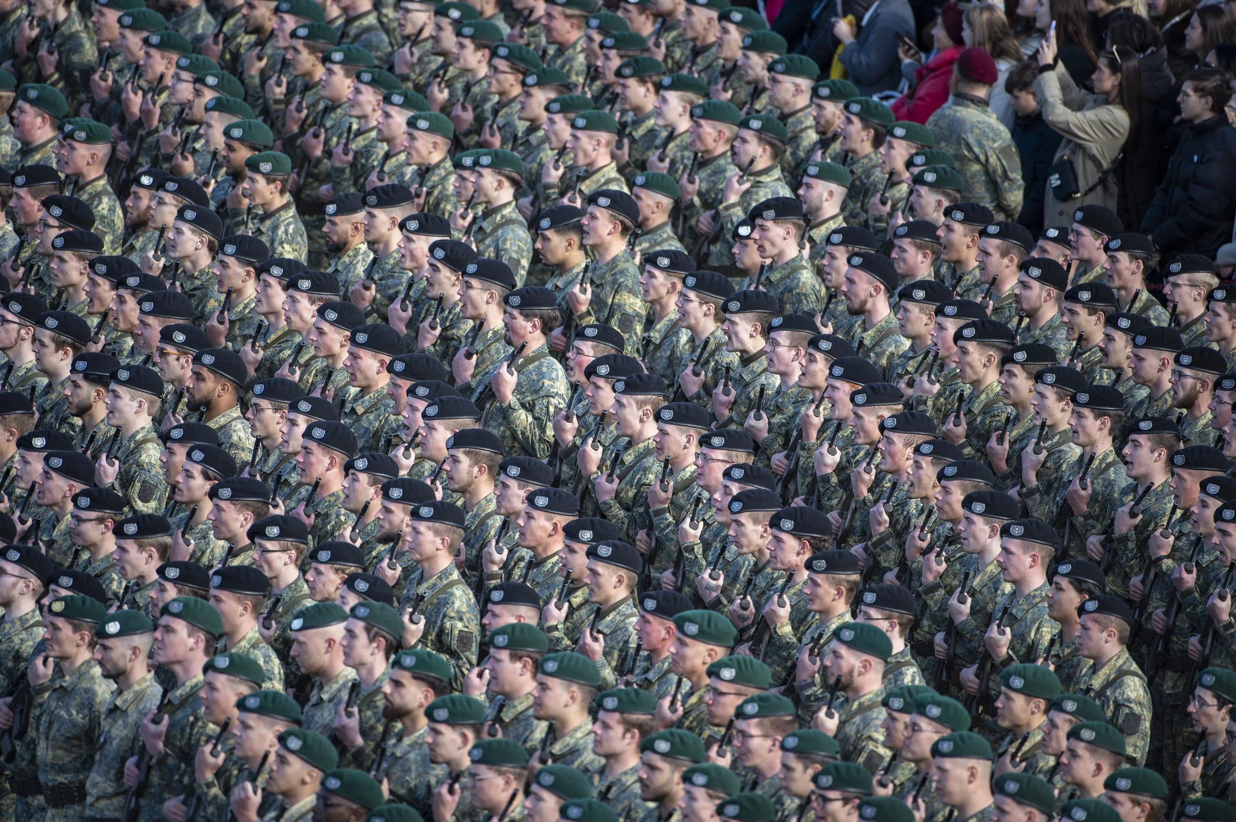 Aufnahme einer großen Gruppe von Soldaten in Uniformen und grünen Mützen, die in einer Parade marschieren, während Zivilpersonen im Hintergrund zuschauen.