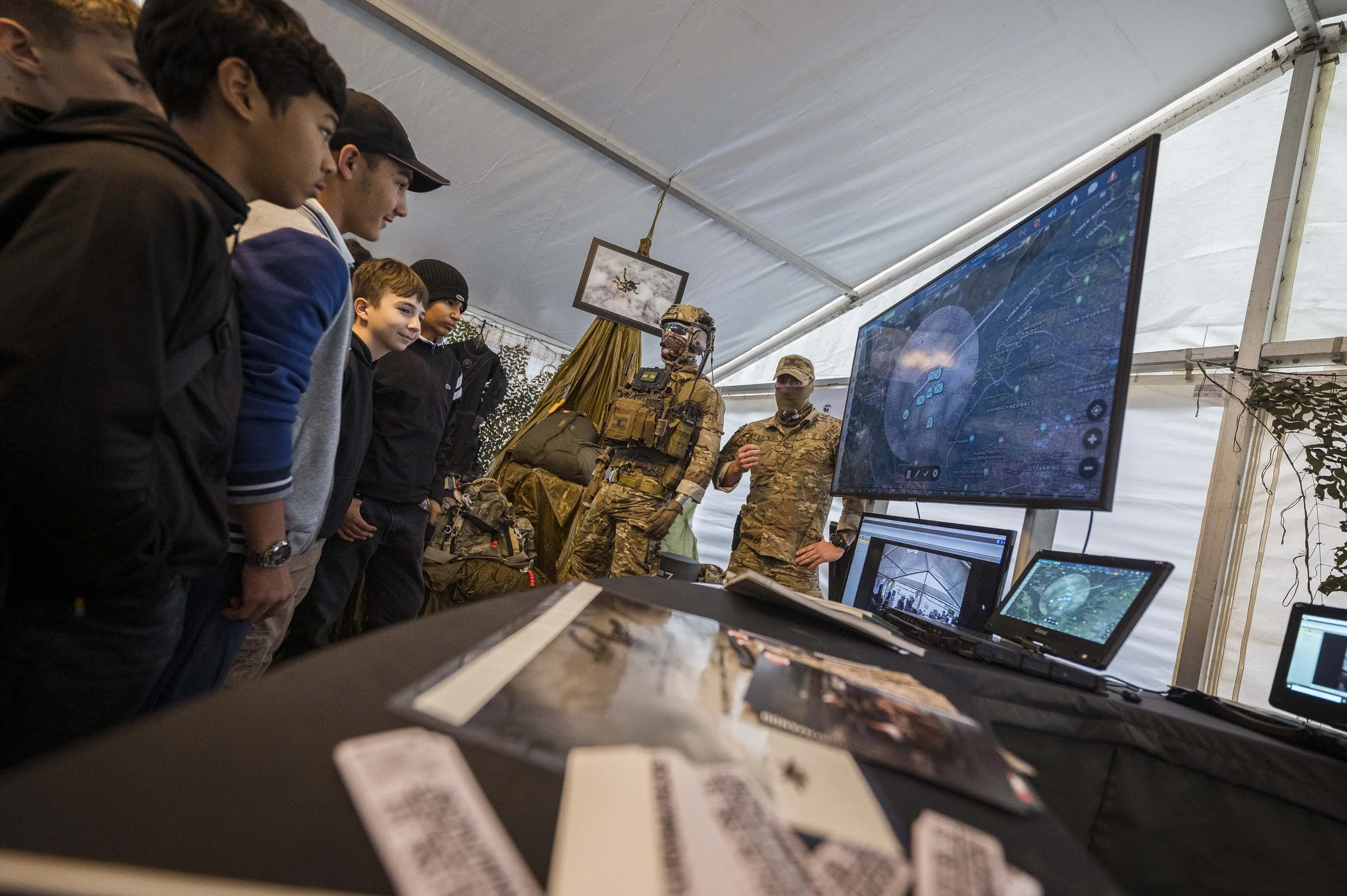 Menschen betrachten eine taktische Demonstration mit Militärpersonal, Computern und Ausrüstung in einem Zelt.