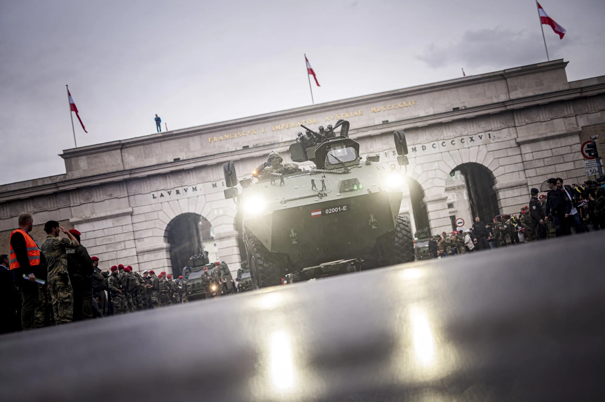 Militärfahrzeug fährt durch ein Tor, umgeben von Menschen und Soldaten, mit einer bewaffneten Person auf dem Fahrzeug, vor einem Gebäude mit lateinischer Inschrift.