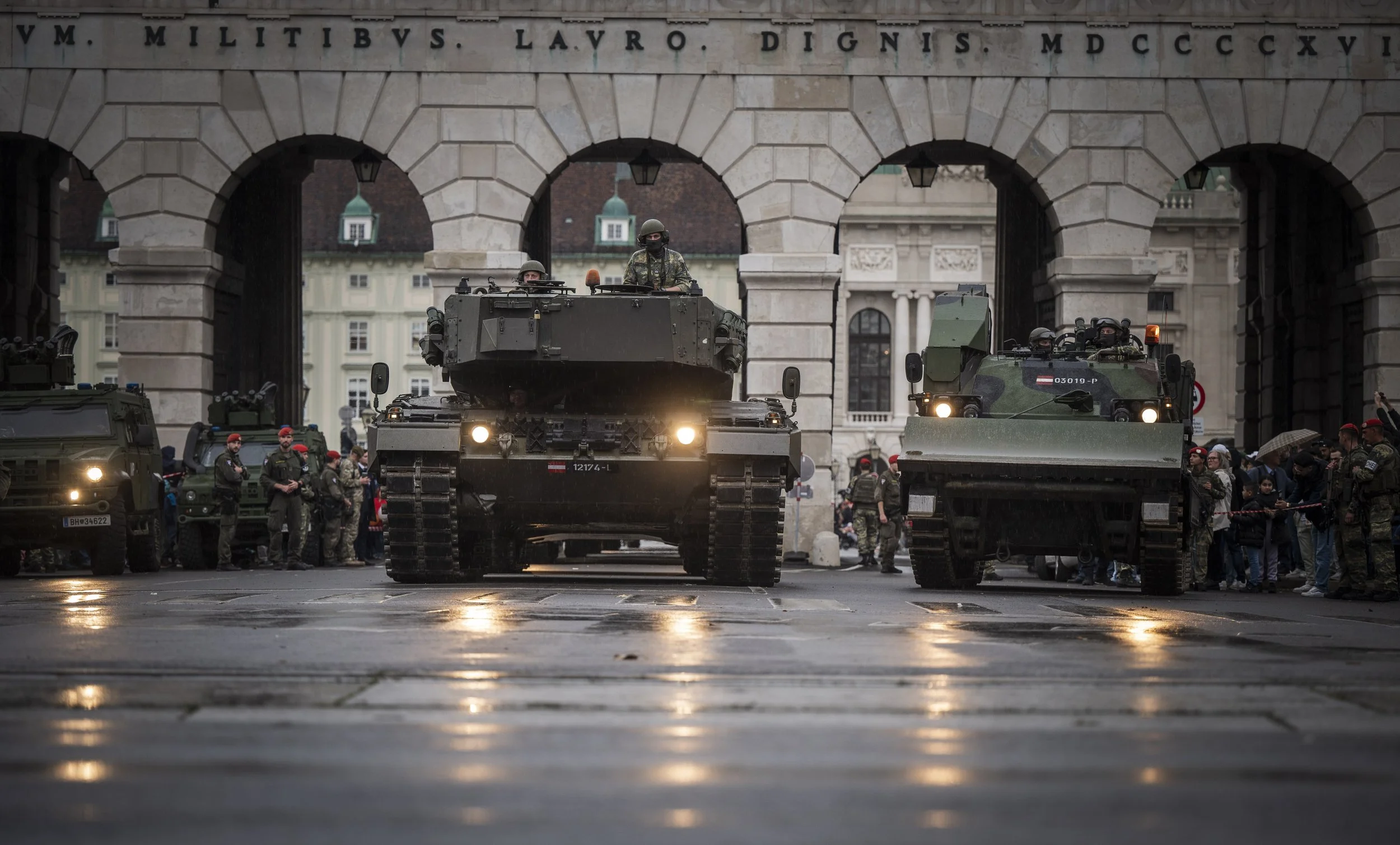 Militärfahrzeuge mit Soldaten und Zuschauern vor einer historischen Stadtmauer, wahrscheinlich bei einer öffentlichen Demonstration oder Parade.