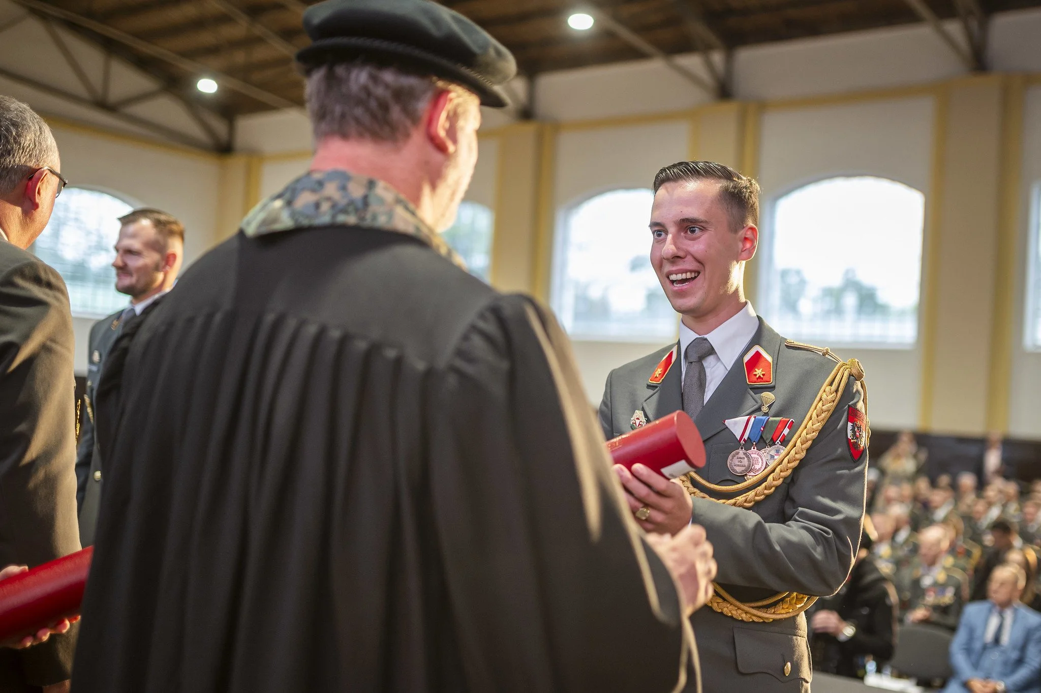 Ein junger Mann in Militäruniform erhält bei einer Zeremonie eine Auszeichnung, umgeben von weiteren Militärangehörigen und Zuschauern.