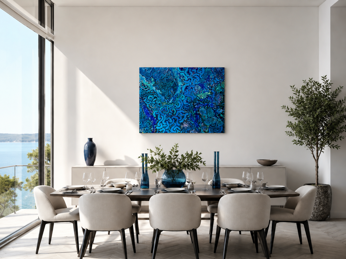 Modern dining room with a large window view of water, decorated with an Indigenous abstract blue artwork, beige chairs, a dark wood table set with white dishes and glasses, and blue vases, greenery, and a potted plant.