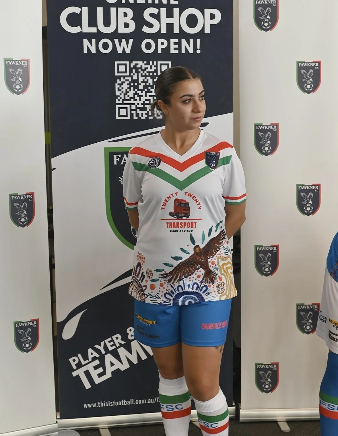 A woman standing in front of a dark blue and white sign that says 'Online Club Shop Now Open!' with a QR code. She is wearing a white sports jersey with red, green, and orange accents, featuring a bird illustration, a red truck logo, and the words 'T