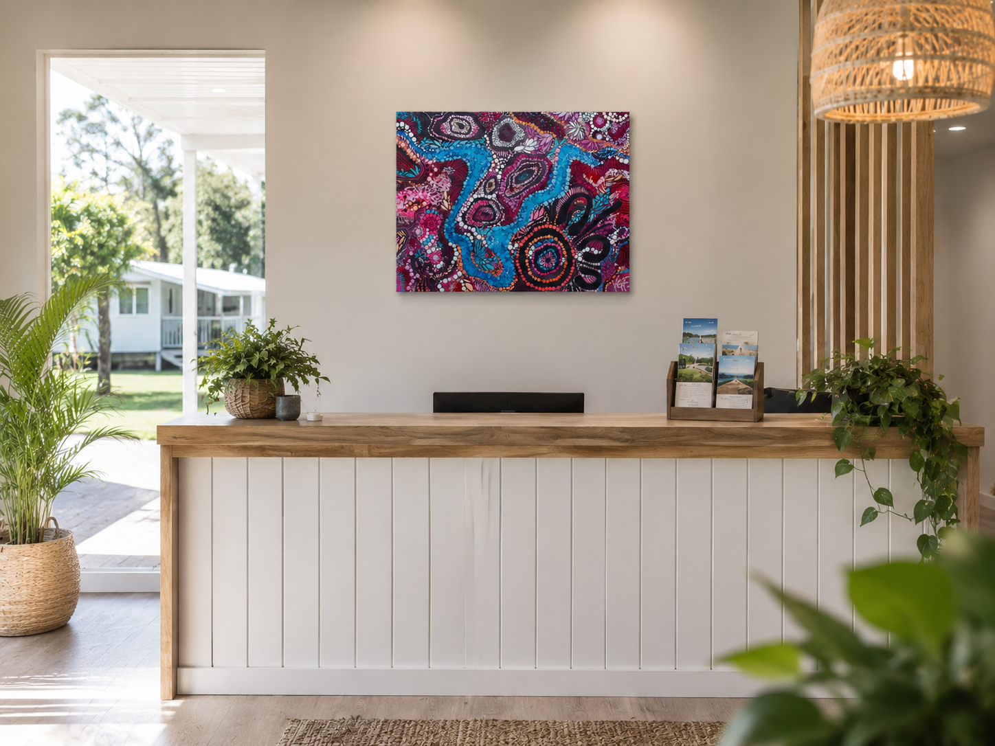 A modern reception desk with plants, colorful Indigenous abstract artwork on the wall, brochure holder, and a wicker pendant light in a bright, welcoming interior space.