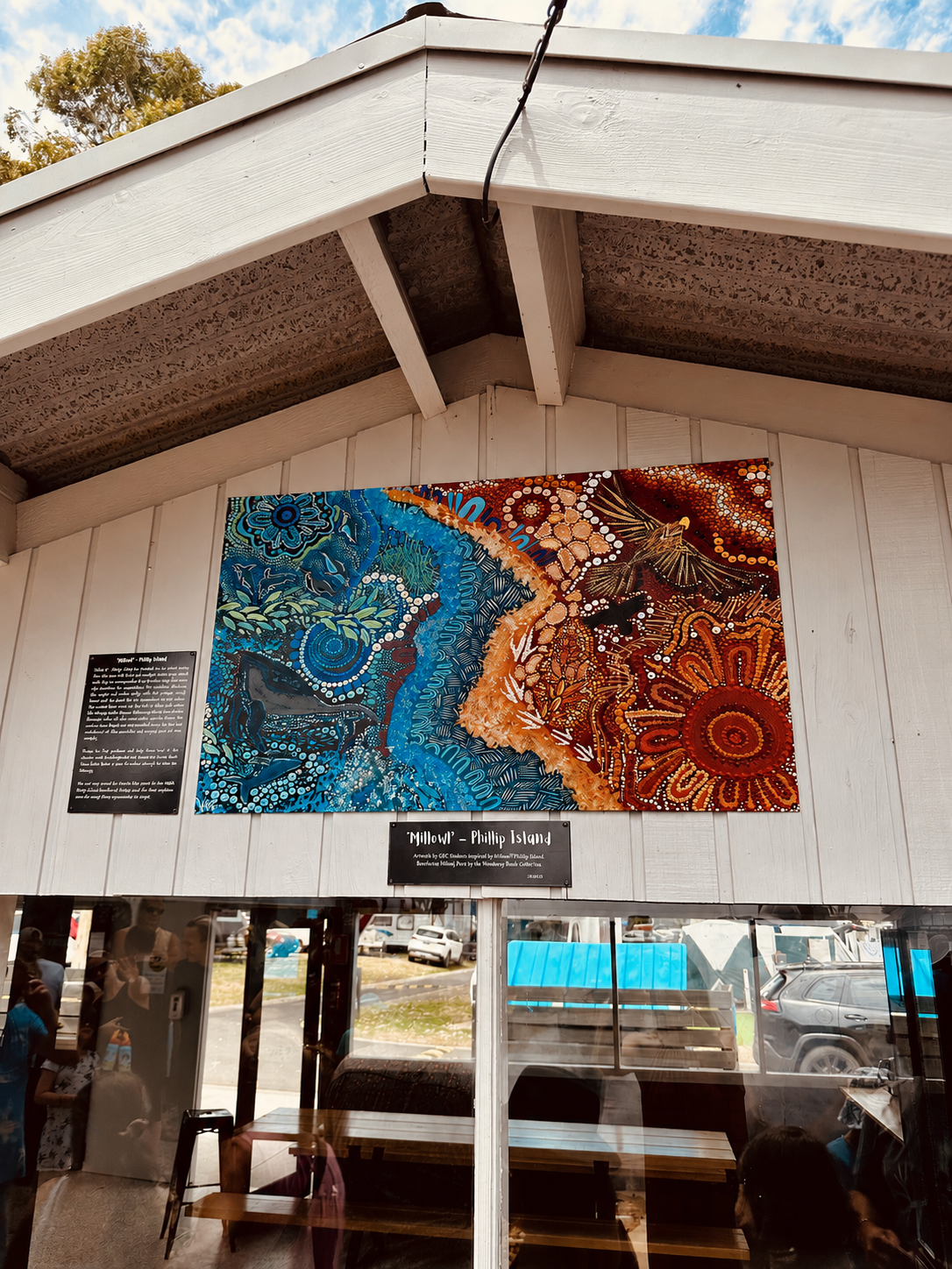 Colorful Aboriginal / Indigenous style painting titled 'Millowi' depicting Phillip Island, featuring vibrant blue, orange, and red patterns with marine and land elements, displayed on a white wooden wall inside a building with glass windows.