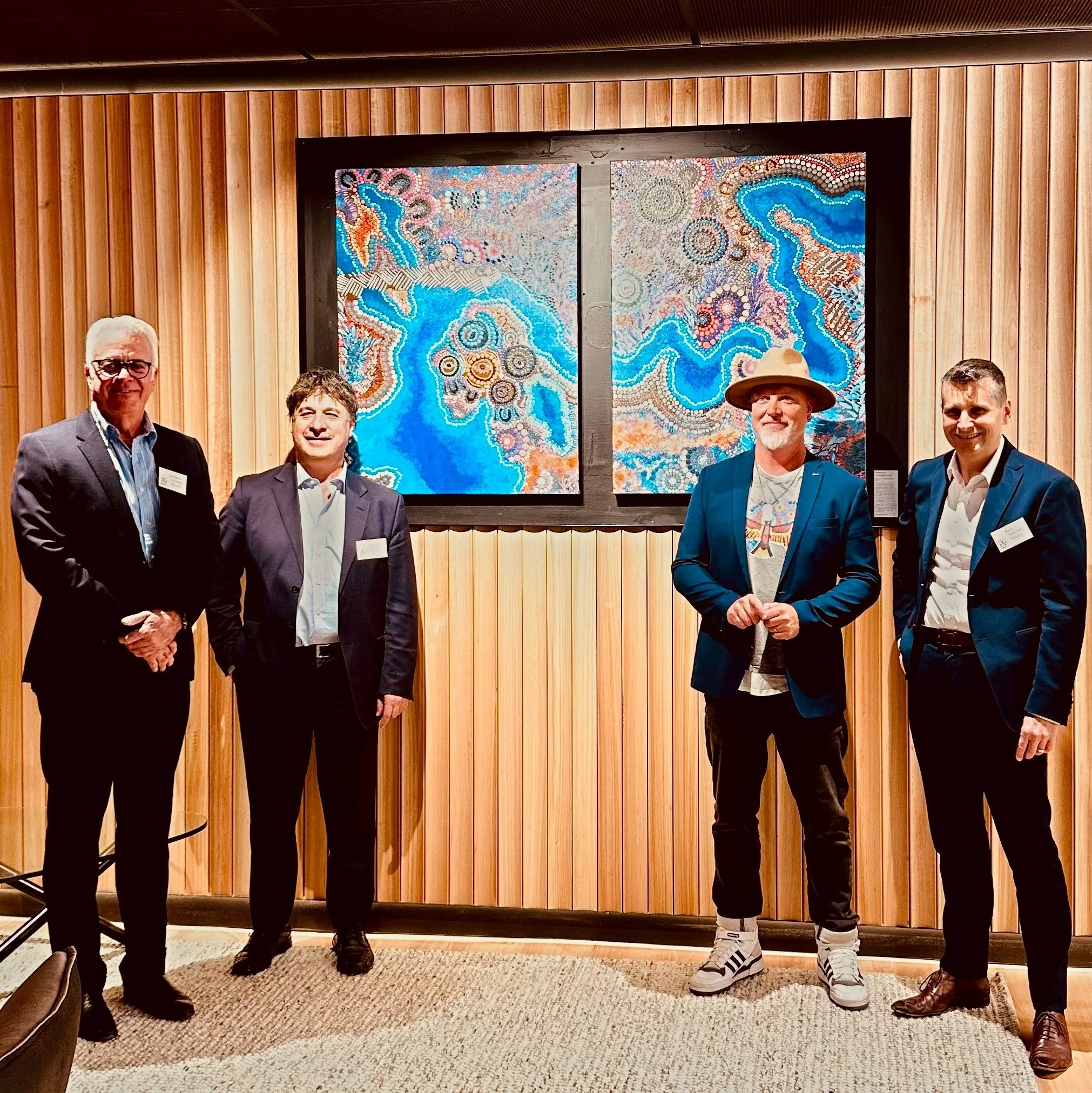 Group of five men standing in front of a colourful Indigenous abstract art piece on a wooden wall, at an indoor event.