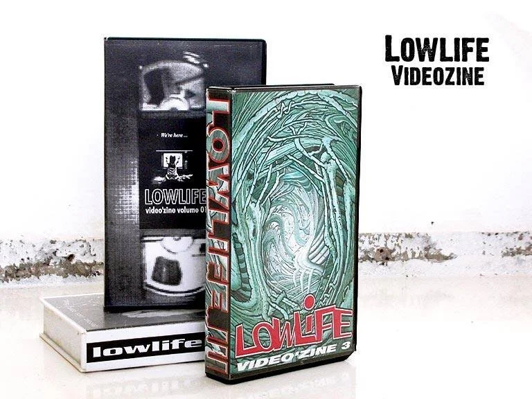Twee VHS-casettes met de labels 'Lowlife Video' en 'Lowlife Videozinine Volume 0', geplaatst op een wit oppervlak tegen een witte muur.