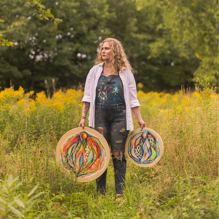Femme avec cheveux bouclés tenant deux tableaux d'art abstrait dans une prairie verte avec des fleurs jaunes et des arbres en arrière-plan.