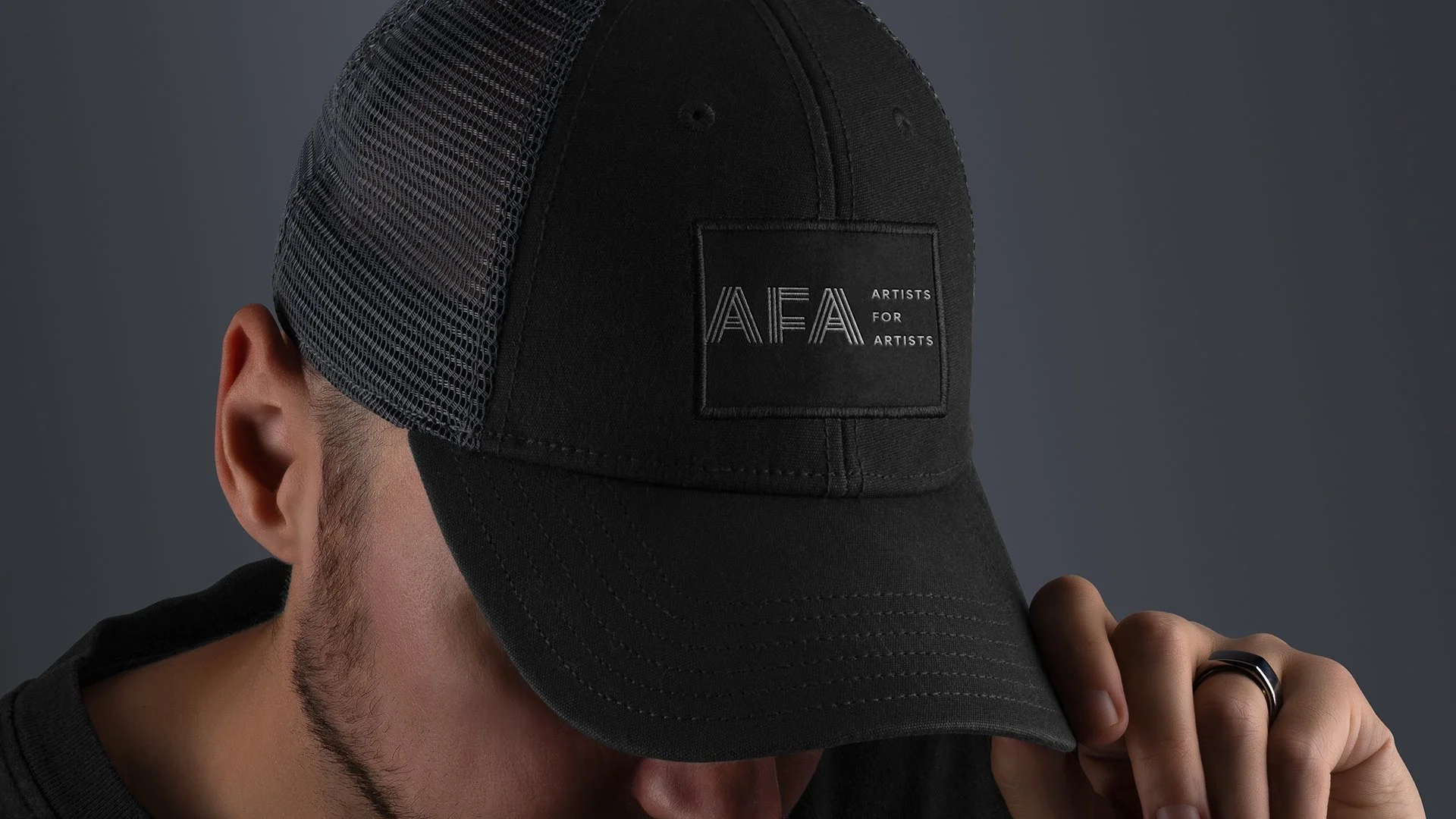 A man wearing a black and gray trucker hat with a patch on the front that reads "AFA: Artists for Artists". He is touching the brim of his hat with his right hand, which has a black ring. His face is partially obscured, and he has light skin and a sh