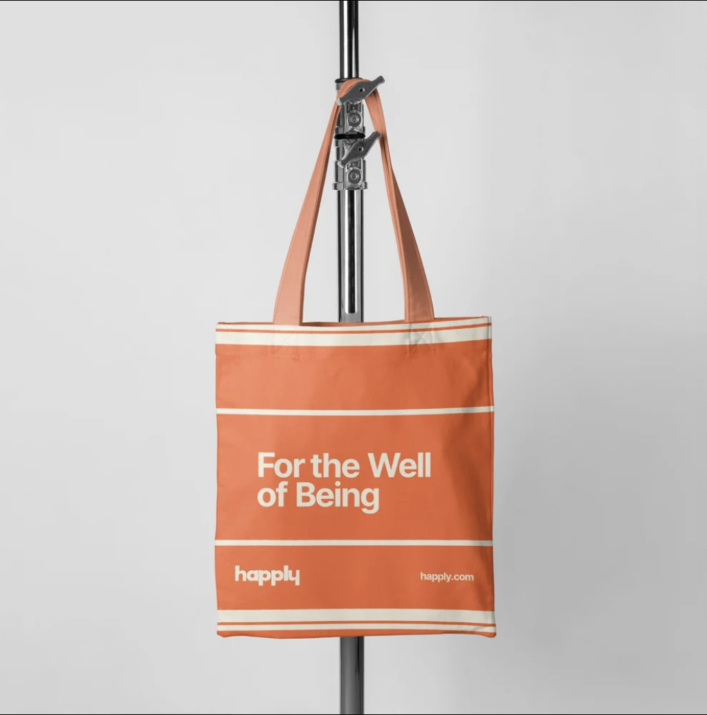 Orange tote bag hanging from a pole with the text 'For the Well of Being' and 'happily' printed on it, against a plain background.