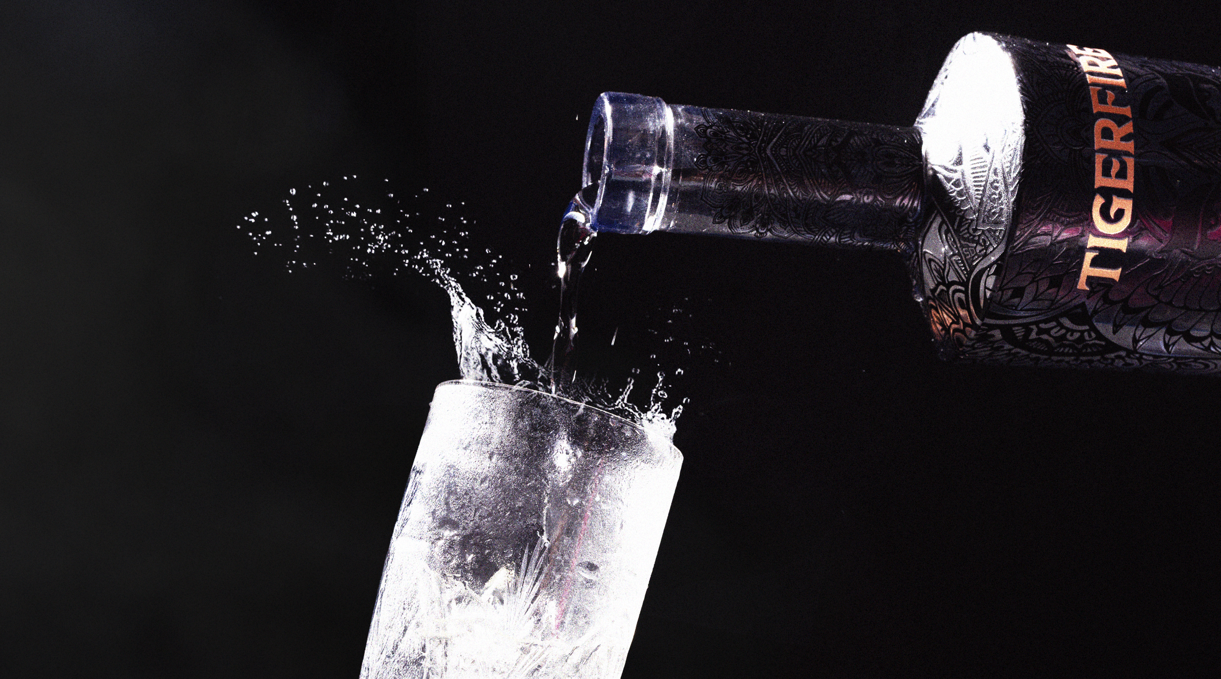 A bottle of white tequila pouring liquid into a tall glass with ice, against a black background.