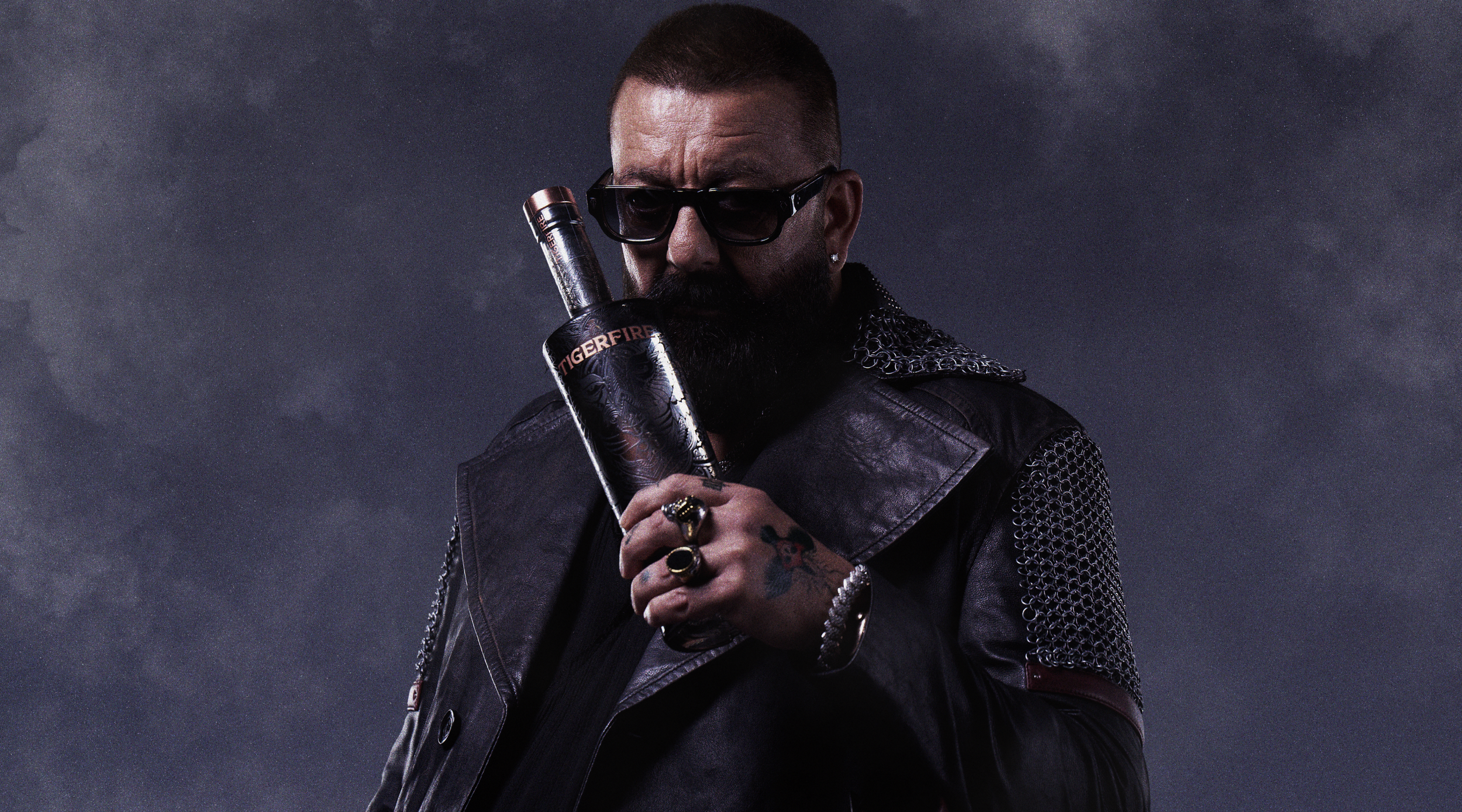 A man with sunglasses and a beard is holding a bottle labeled 'Tigerfire' to his lips. He is dressed in black leather jacket with chainmail details and has tattoos and rings on his fingers. The background is dark and cloudy.