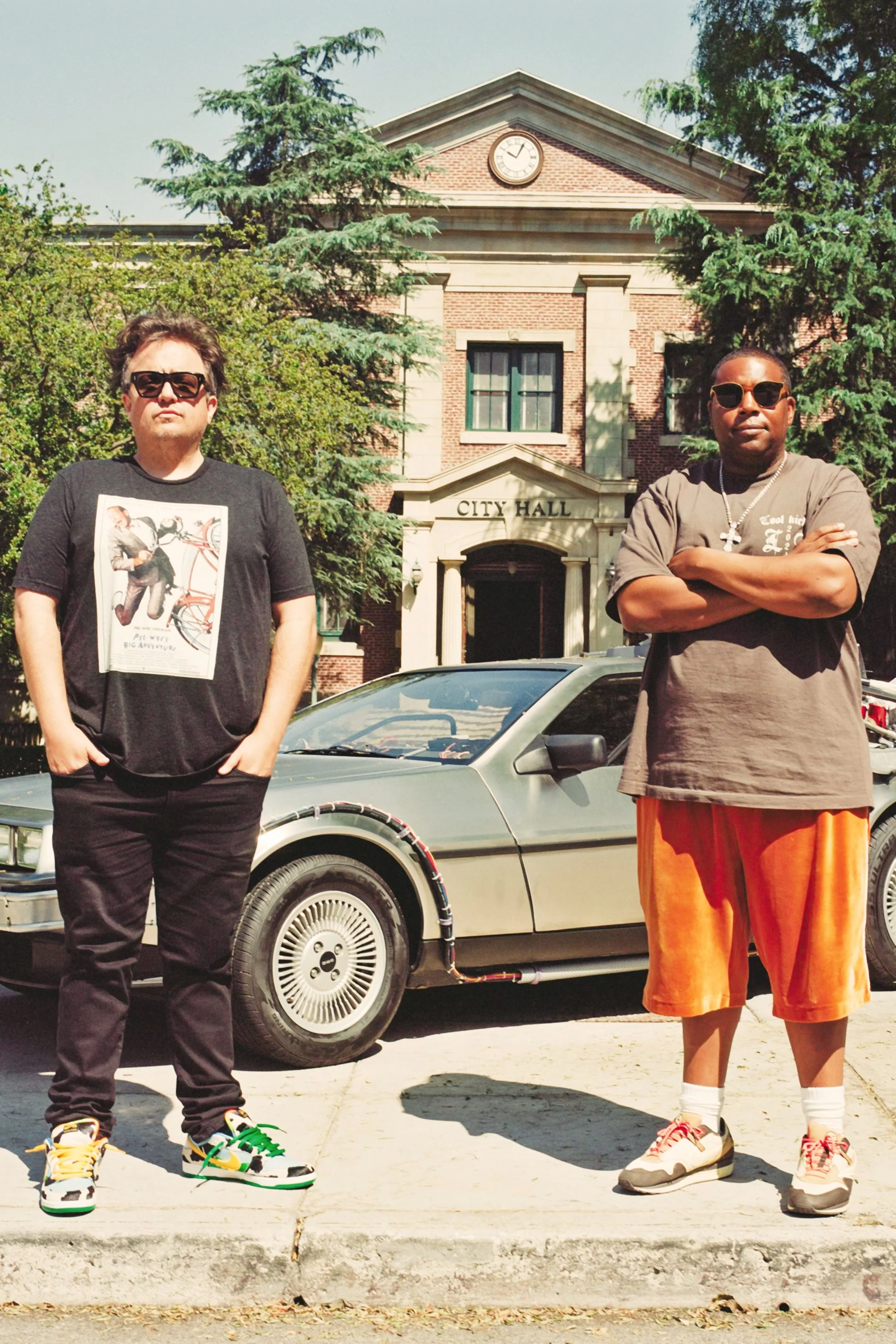 Two men standing in front of a vintage car and a city hall building, with trees behind them on a sunny day.