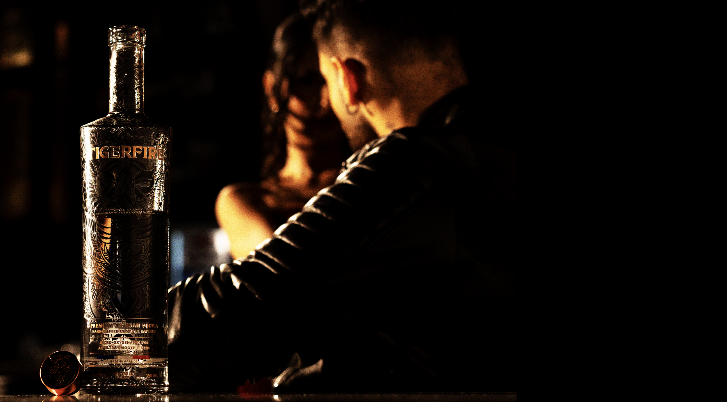 A bottle of Tiger Fire premium artisan vodka on a table with a cap next to it, with a couple in the background sitting close and facing each other in a dimly lit setting.