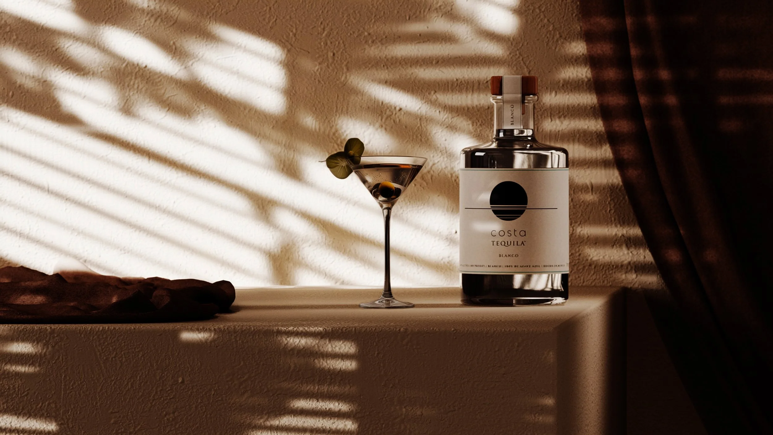 A bottle of Costa Tequila Blanco, a martini glass with an olive and garnish, on a beige surface with curtains casting striped sunlight shadows on a textured beige wall.