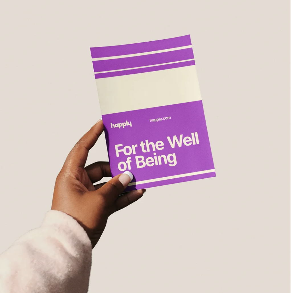 A hand holding a purple and white box with the brand name happily and the phrase "For the Well of Being" on it.