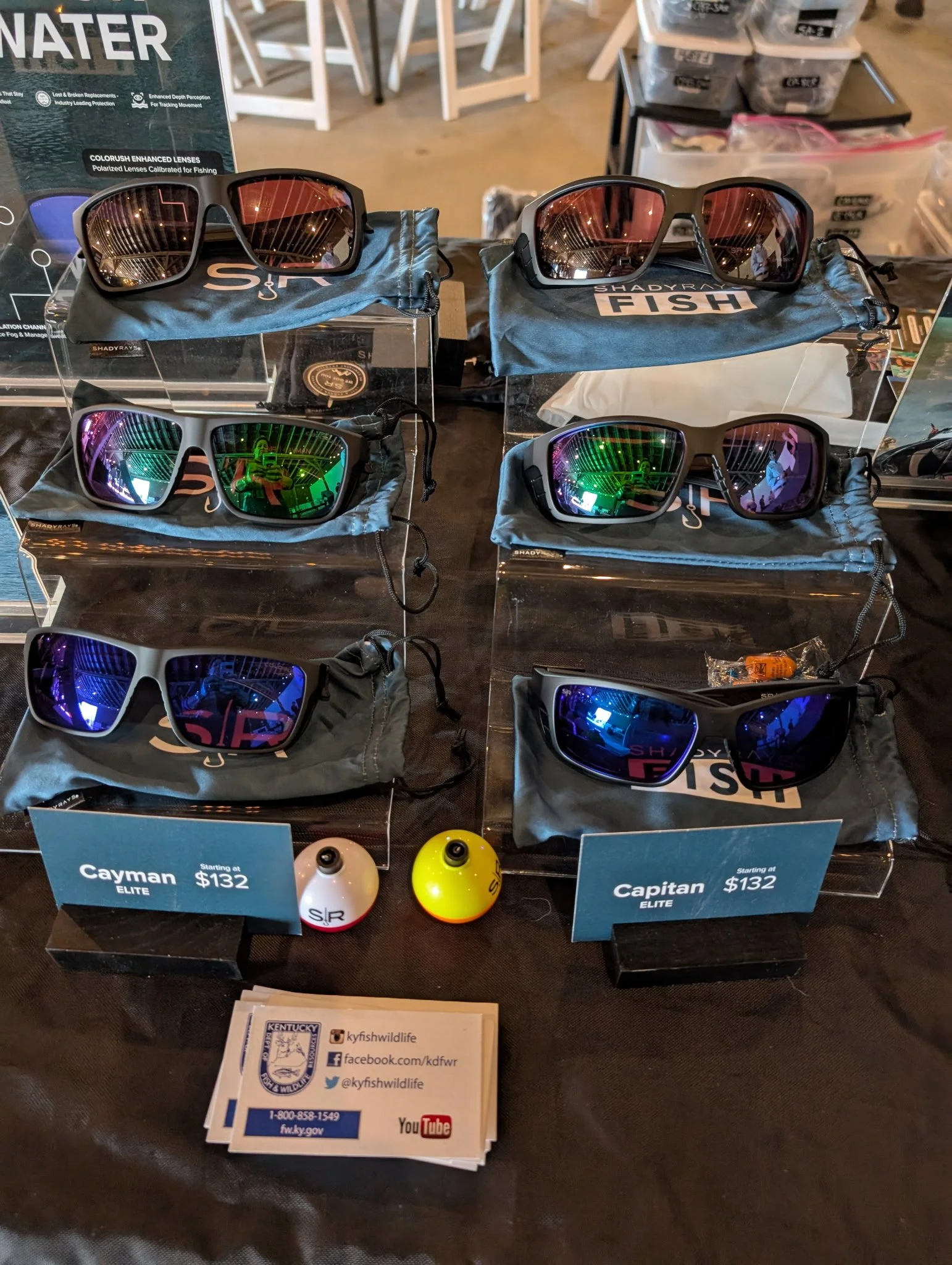 Display of sunglasses and fishing gear, including six pairs of sunglasses with colored reflective lenses, two spherical fishing floats, and informational signs pricing starting at $132, with branding and informational materials about Kentucky Fish and Wildlife.