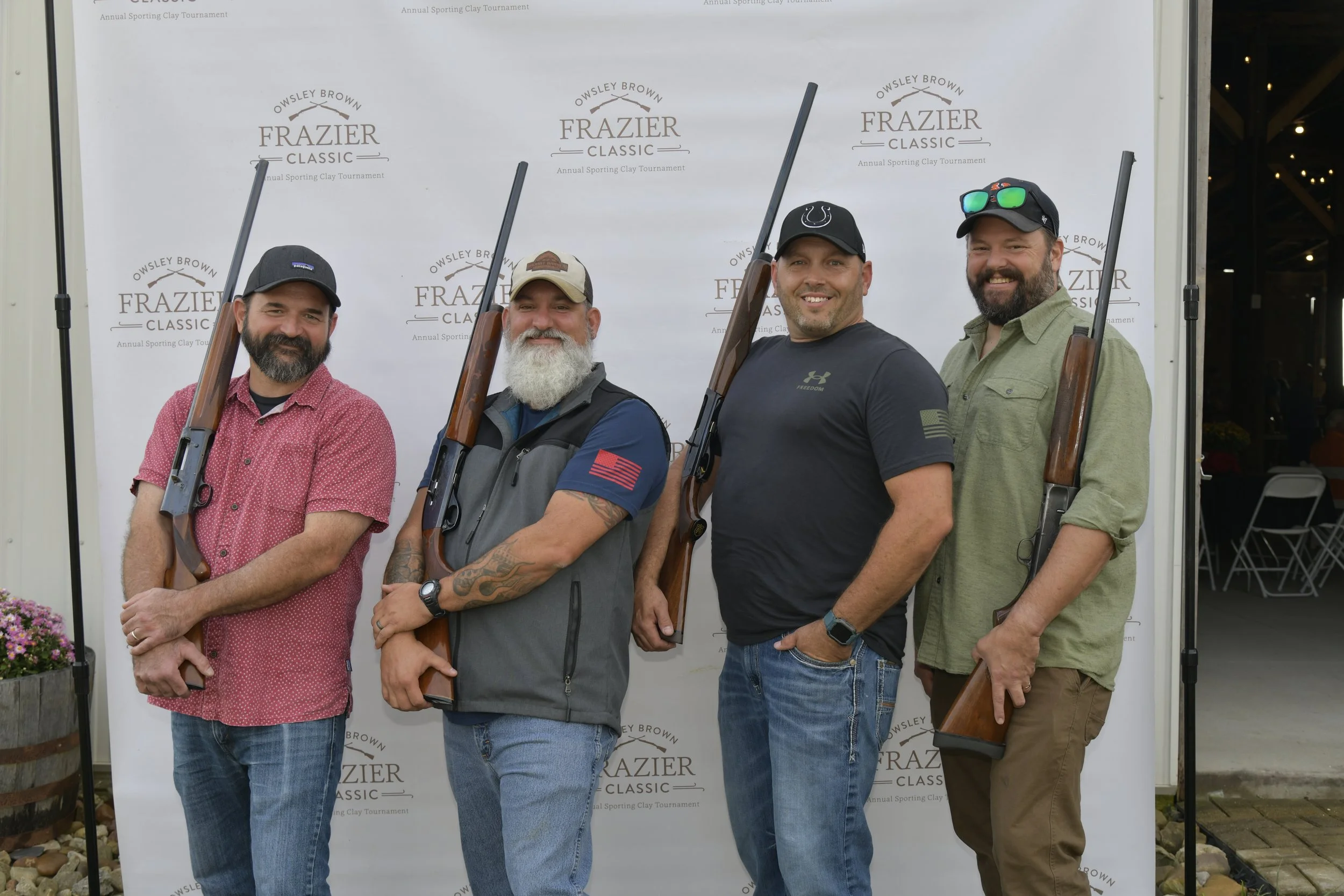 Four men standing outdoors holding rifles at the Frazier Classic annual sporting clay tournament, posing in front of a backdrop.