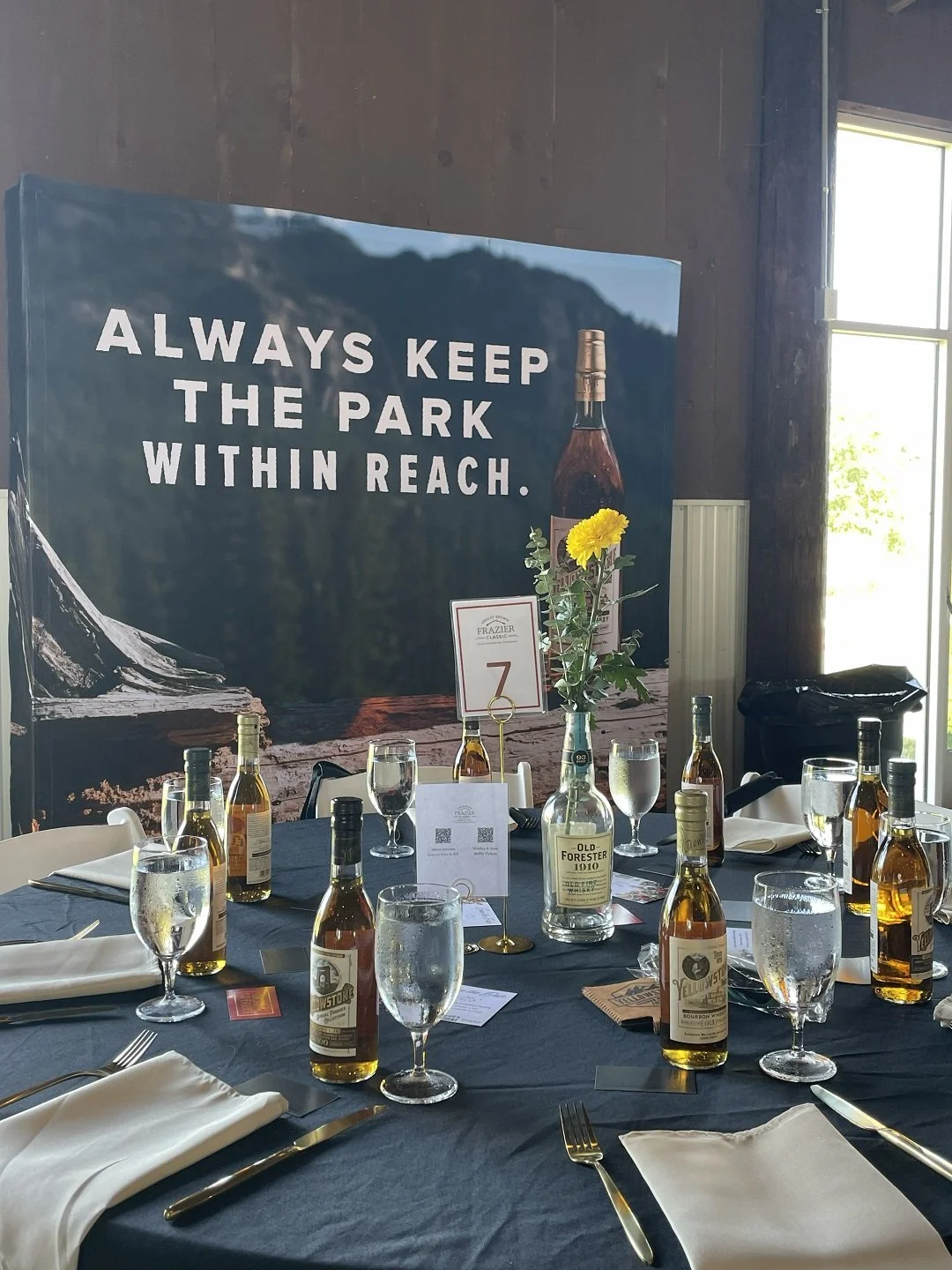 Indoor banquet table with white napkins, silverware, multiple bottles of whiskey, glasses filled with water, a flower vase with yellow flowers, and a large wall sign that says, "Always keep the park within reach".