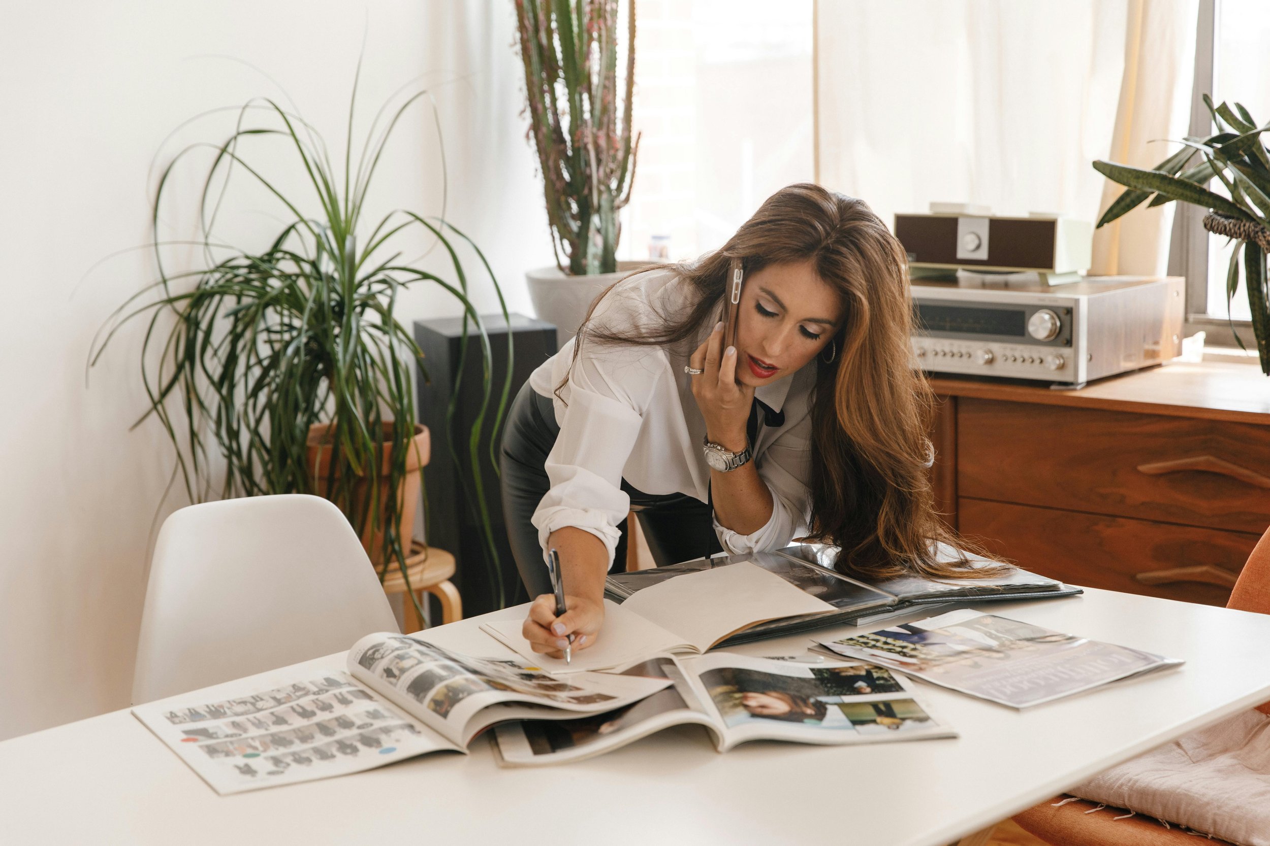 A woman with long brown hair is standing at a table, looking at photo albums and writing in one. She is wearing a white blouse and black leather pants, talking on a cellphone, and appears to be focused.