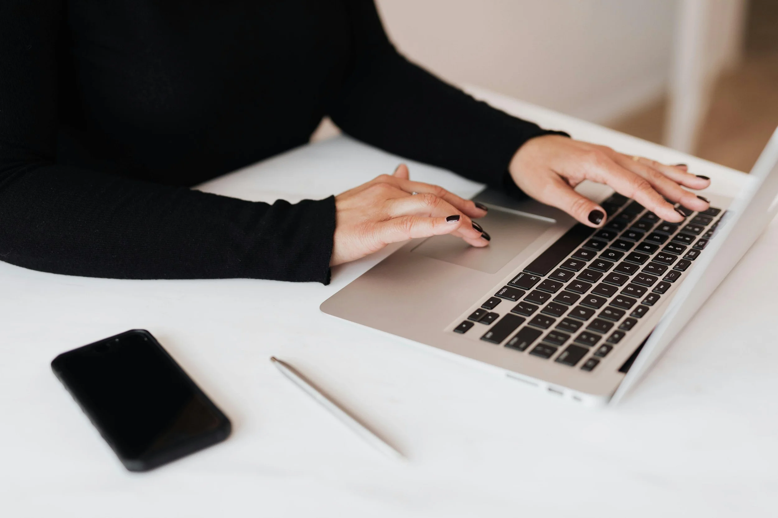 Person in black long-sleeve shirt typing on a silver laptop on a white desk, with a black smartphone and a white pen nearby.