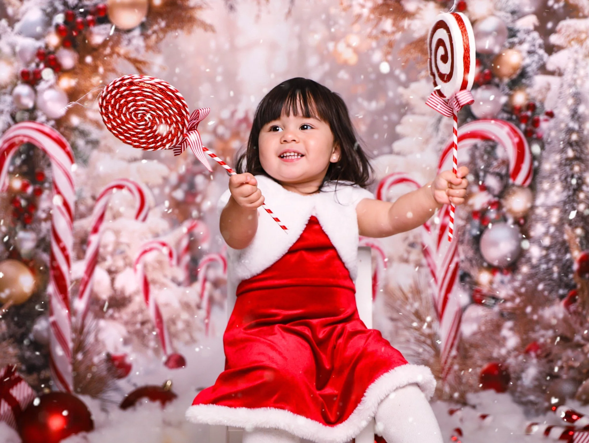 A young girl dressed in a red Santa dress with white fur trim, sitting in front of a decorated Christmas tree, holding candy canes and smiling.