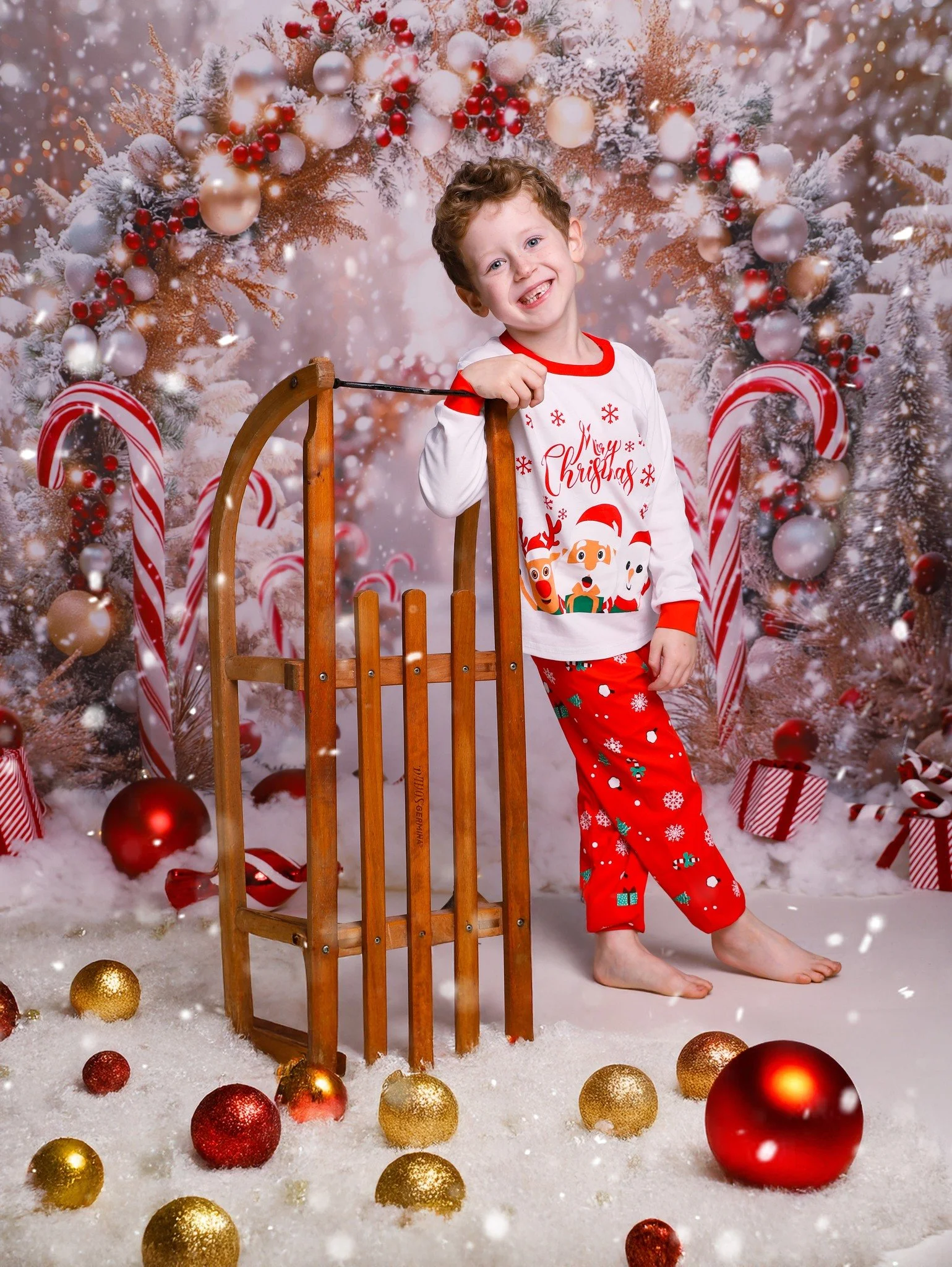 A young boy in Christmas pajamas stands indoors with a wooden sled, surrounded by Christmas decorations and ornaments, in front of a festive holiday background with snow, candy canes, and ornaments.
