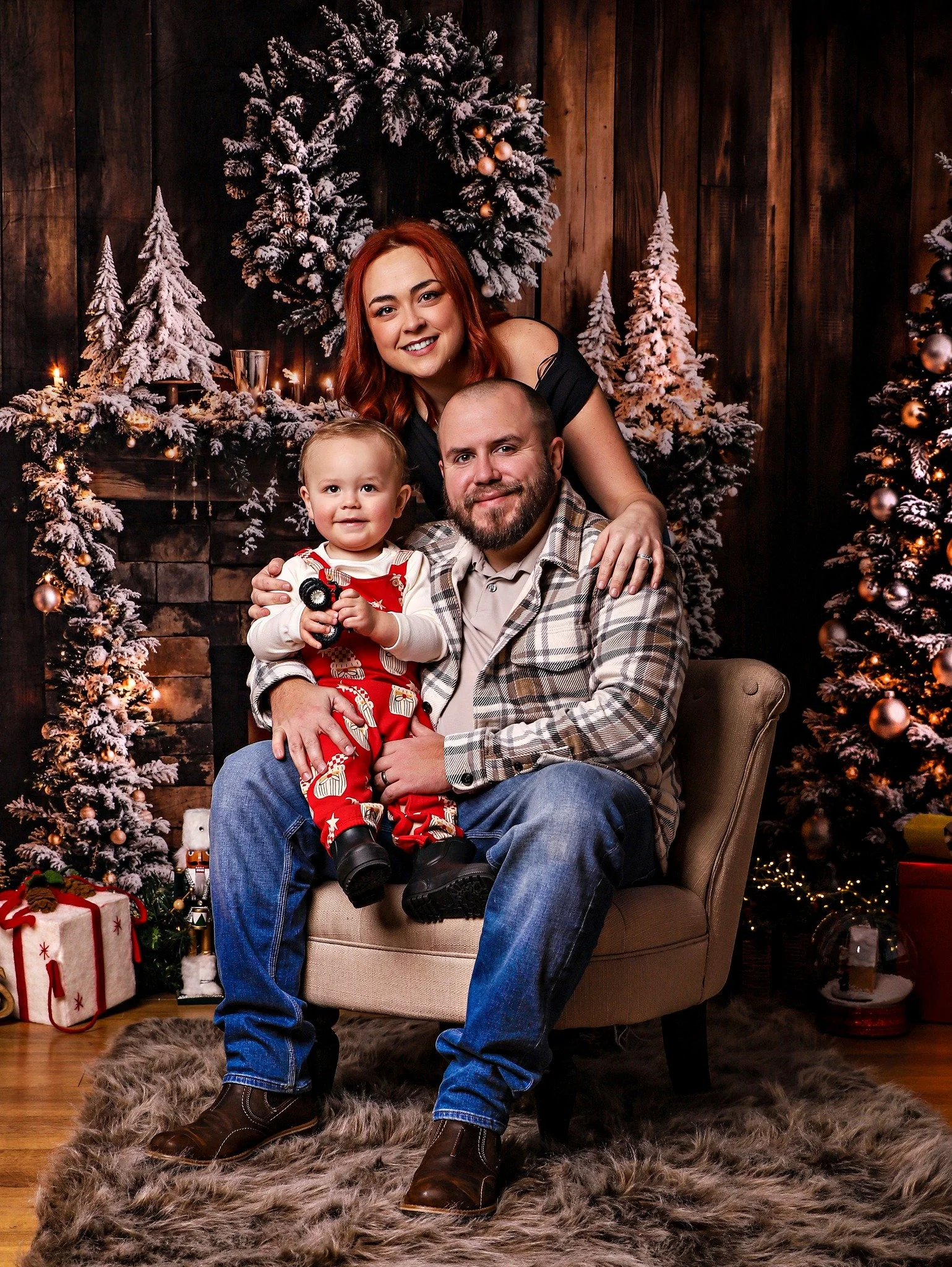 A family of three, including a woman with red hair, a man with a beard, and a small child with blonde hair, pose at Christmas with snow-dusted Christmas trees and decorations in the background.