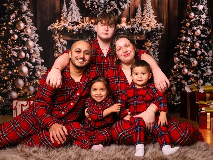 Family of five in matching red plaid pajamas sitting in front of decorated Christmas trees with wrapped gifts.