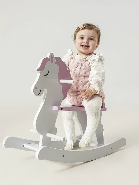 A smiling young girl sitting on a pink and white rocking horse toy against a plain background from their cake smash photoshoot at Meadowside Studio in Warrington to celebrate their 1st birthday!