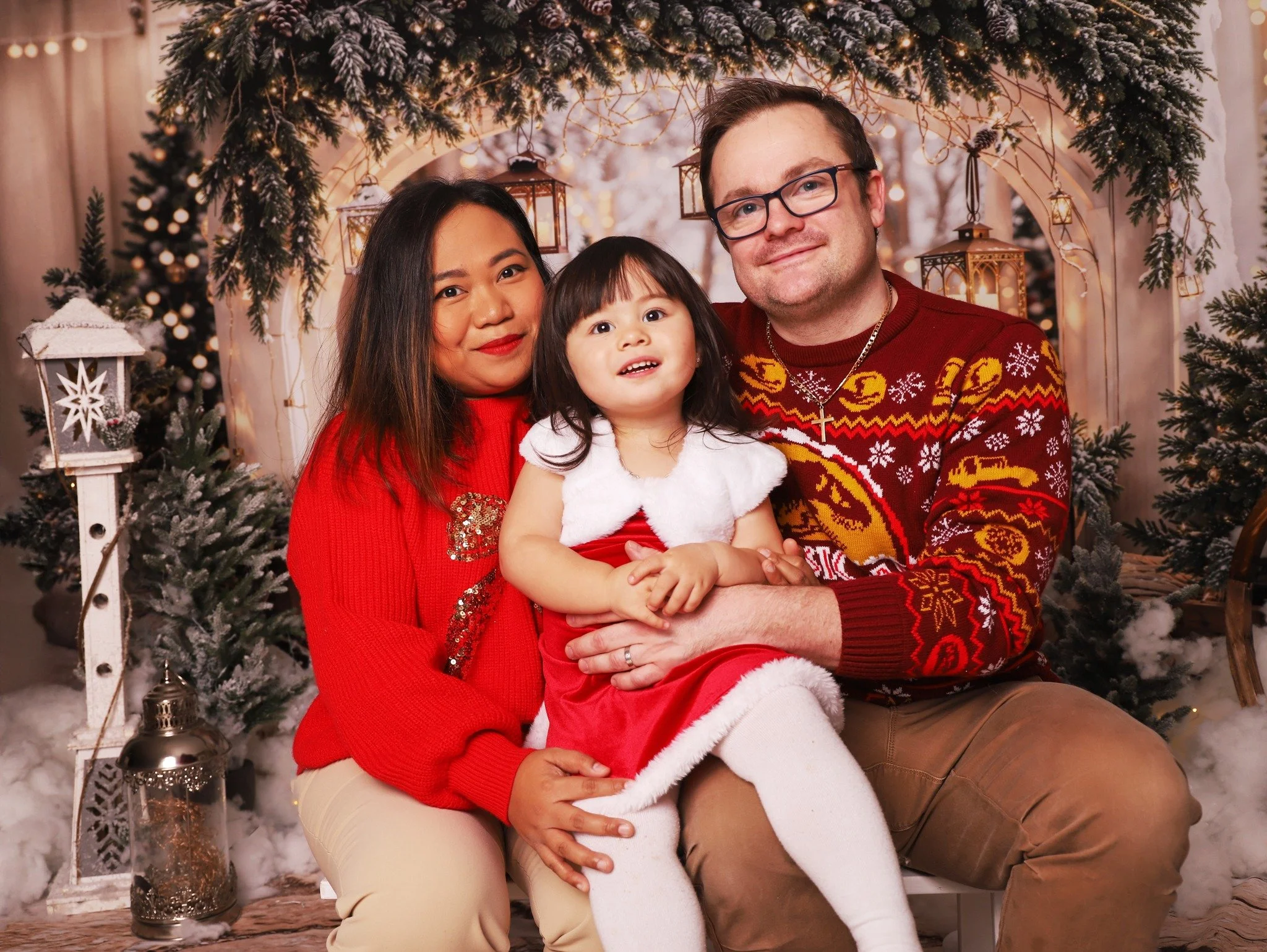 Family of three sitting together in holiday-themed setting. Woman in red sweater, man in Christmas sweater, young girl in Santa dress, surrounded by Christmas trees, lanterns, and twinkling lights.