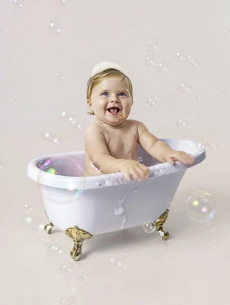 A happy baby sitting in a white toy bathtub with golden clawfoot legs, surrounded by floating soap bubbles from their cake smash photoshoot at Meadowside Studio in Warrington to celebrate their 1st birthday!