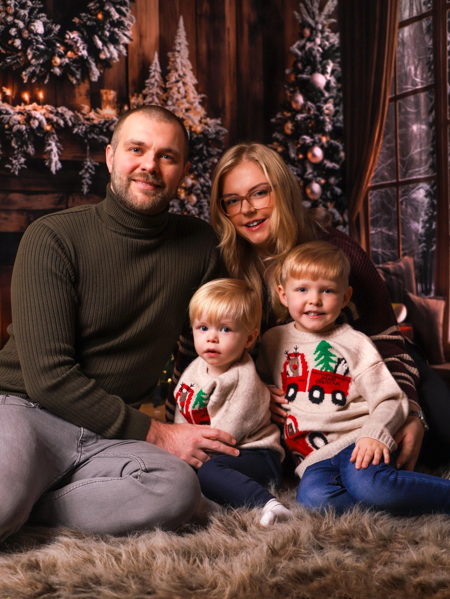 Family of four sitting on a fluffy rug with Christmas sweaters, wooden walls, decorated Christmas trees, and holiday decor in the background.