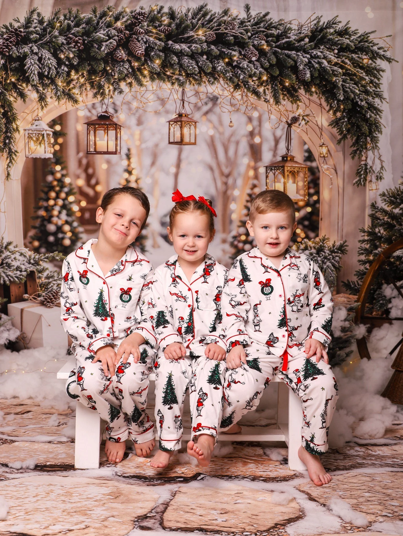 Three children in Christmas pajamas sitting on a white bench in front of a festive snow-covered Christmas scene with decorated trees, lanterns, and snow.