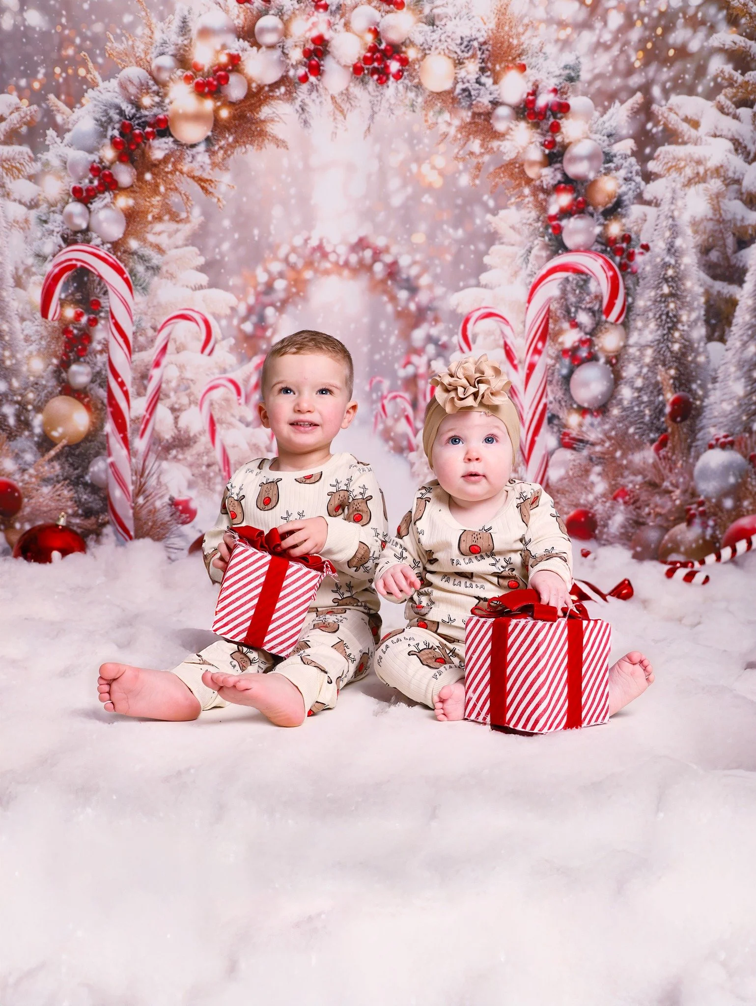 Two children in Christmas pajamas sitting on fake snow, holding striped gift boxes, in front of a decorated Christmas tree with ornaments and candy canes.