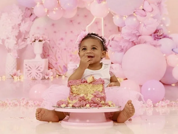 A smiling baby girl sitting on the floor surrounded by pink decorations, with a large slice of pink birthday cake in front of her from their cake smash photoshoot at Meadowside Studio in Warrington to celebrate their 1st birthday!
